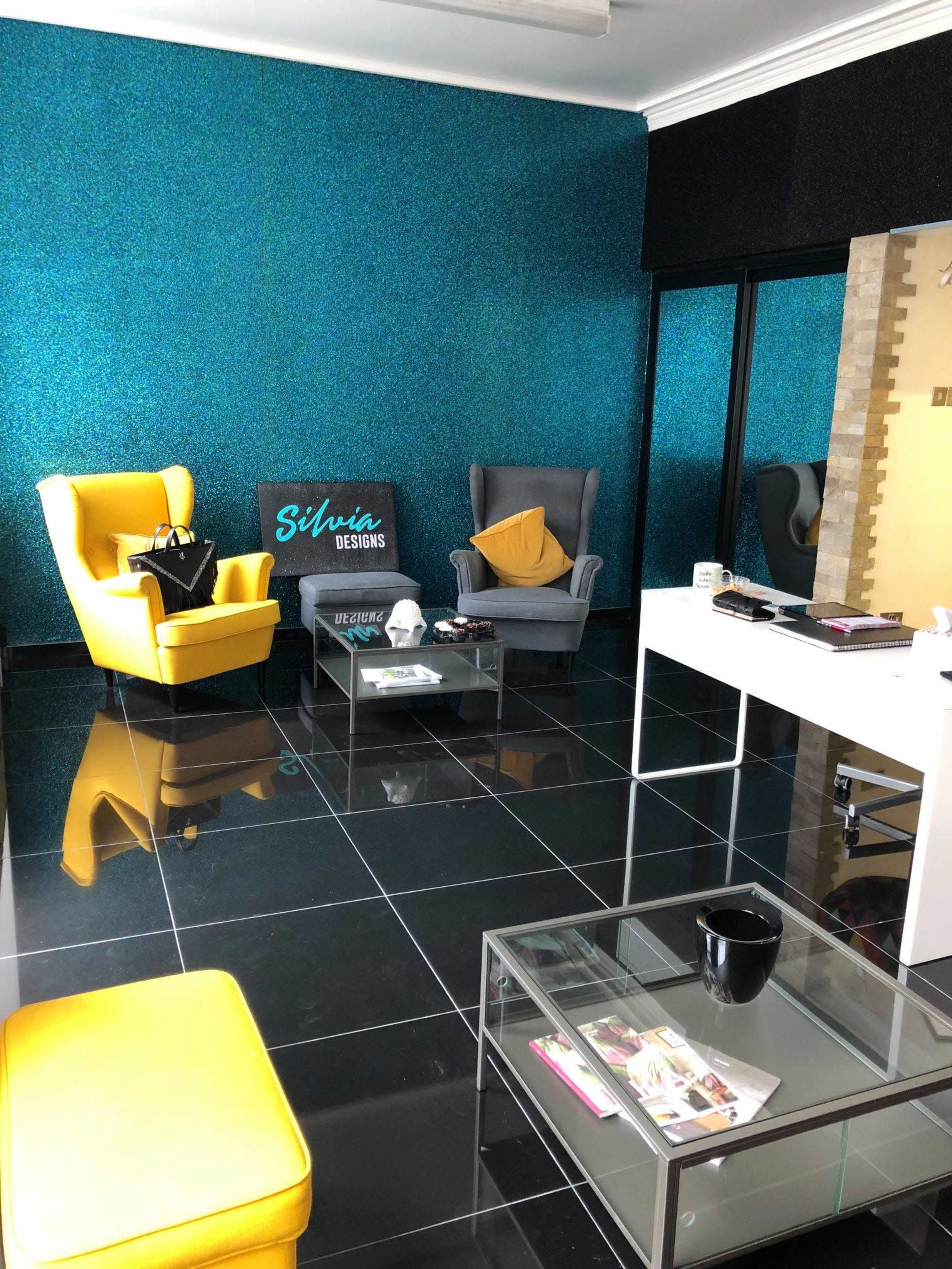 A modern office space featuring a yellow armchair, a gray armchair, a glass coffee table, and a desk against a glittering blue wall.