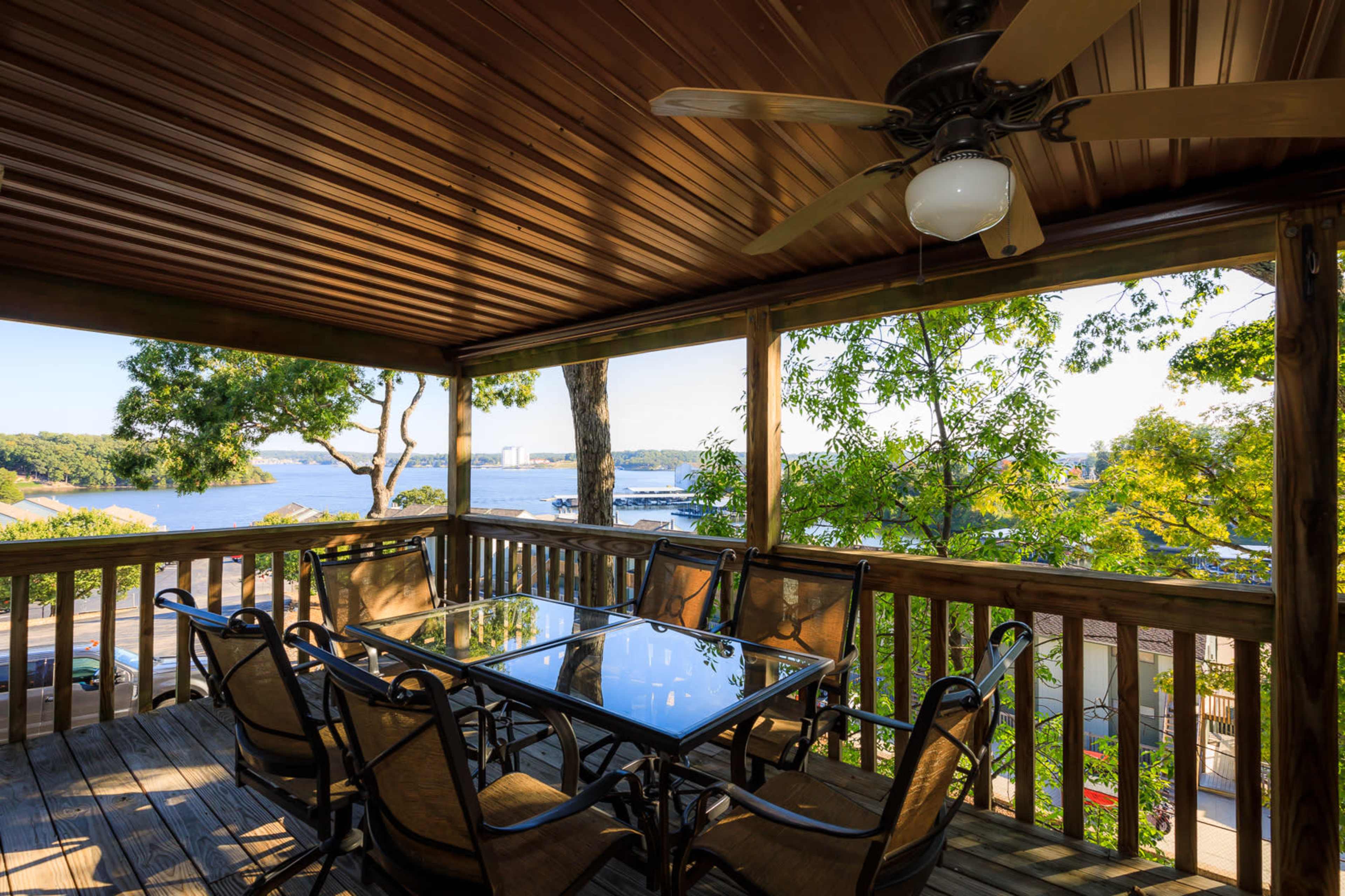 The image shows a covered outdoor dining area with a glass table and chairs, overlooking a body of water and trees.
