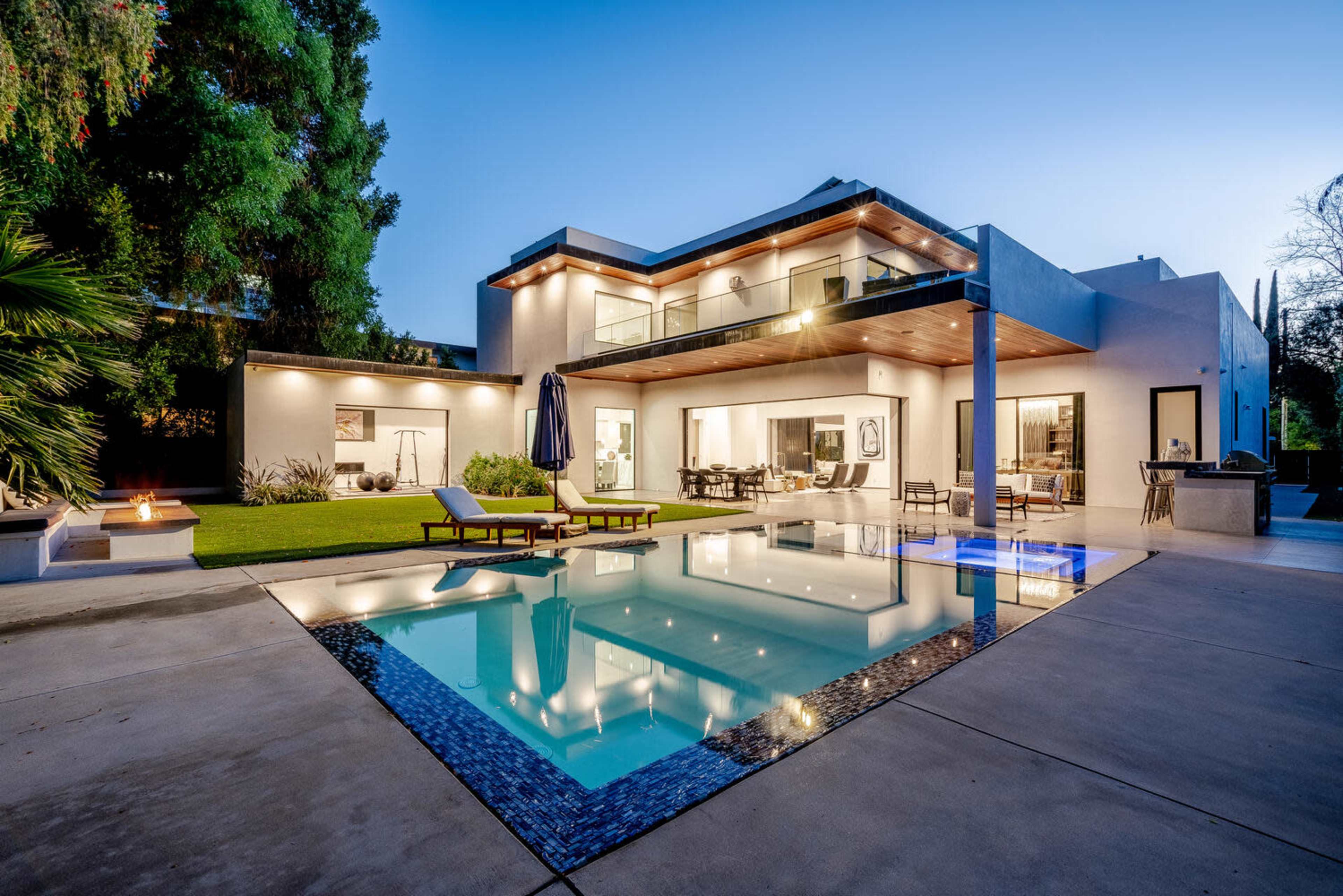 A modern two-story house with a swimming pool and outdoor seating area, surrounded by landscaping and illuminated by evening light.