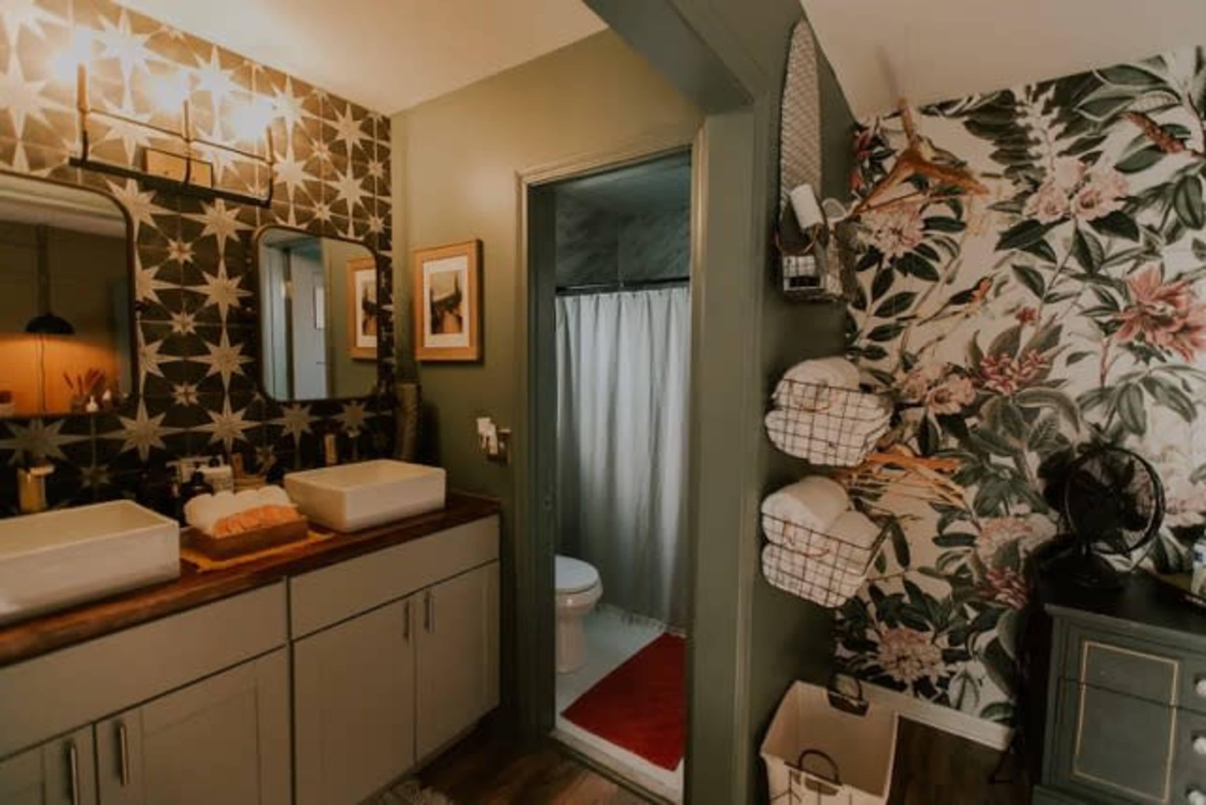 A modern bathroom with green and floral wallpaper, dual sinks, a wooden countertop, and a toilet in a separate area behind a curtain.