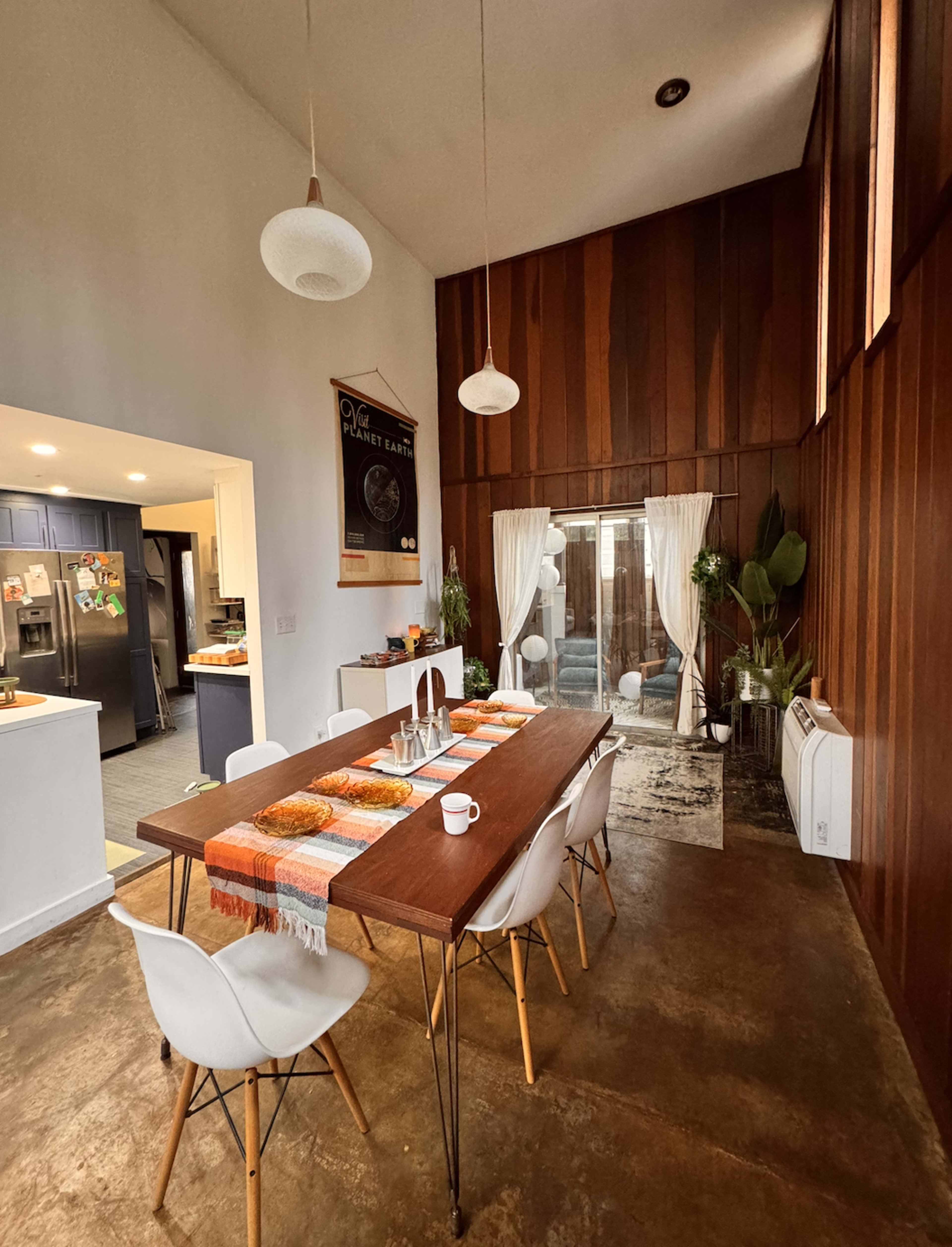 The image shows a modern dining area with a wooden table surrounded by white chairs, plants in the corners, and large windows covered by sheer curtains.