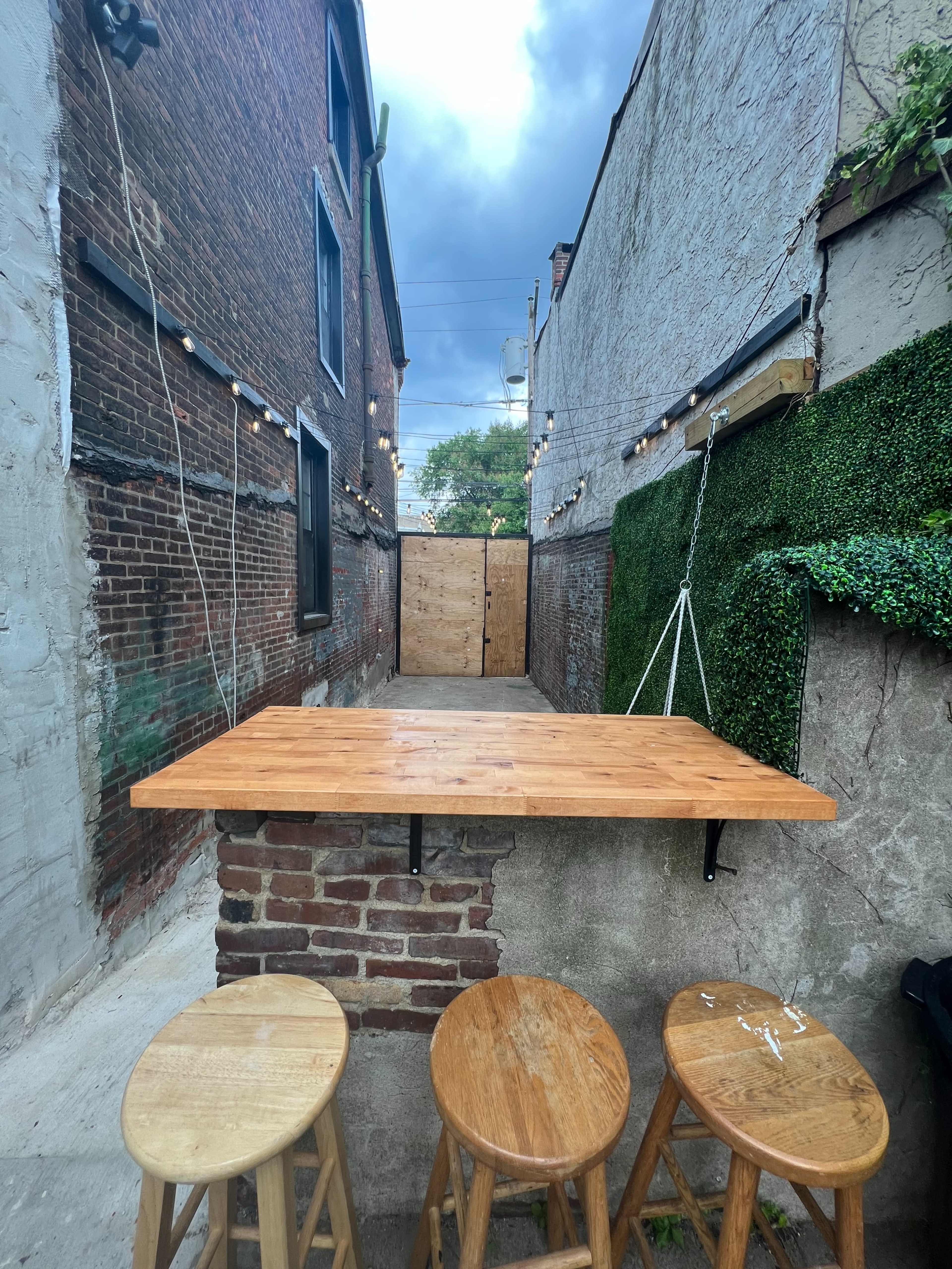 A wooden bar table with three stools is positioned in a narrow alley, flanked by brick and concrete walls, with greenery and string lights overhead.