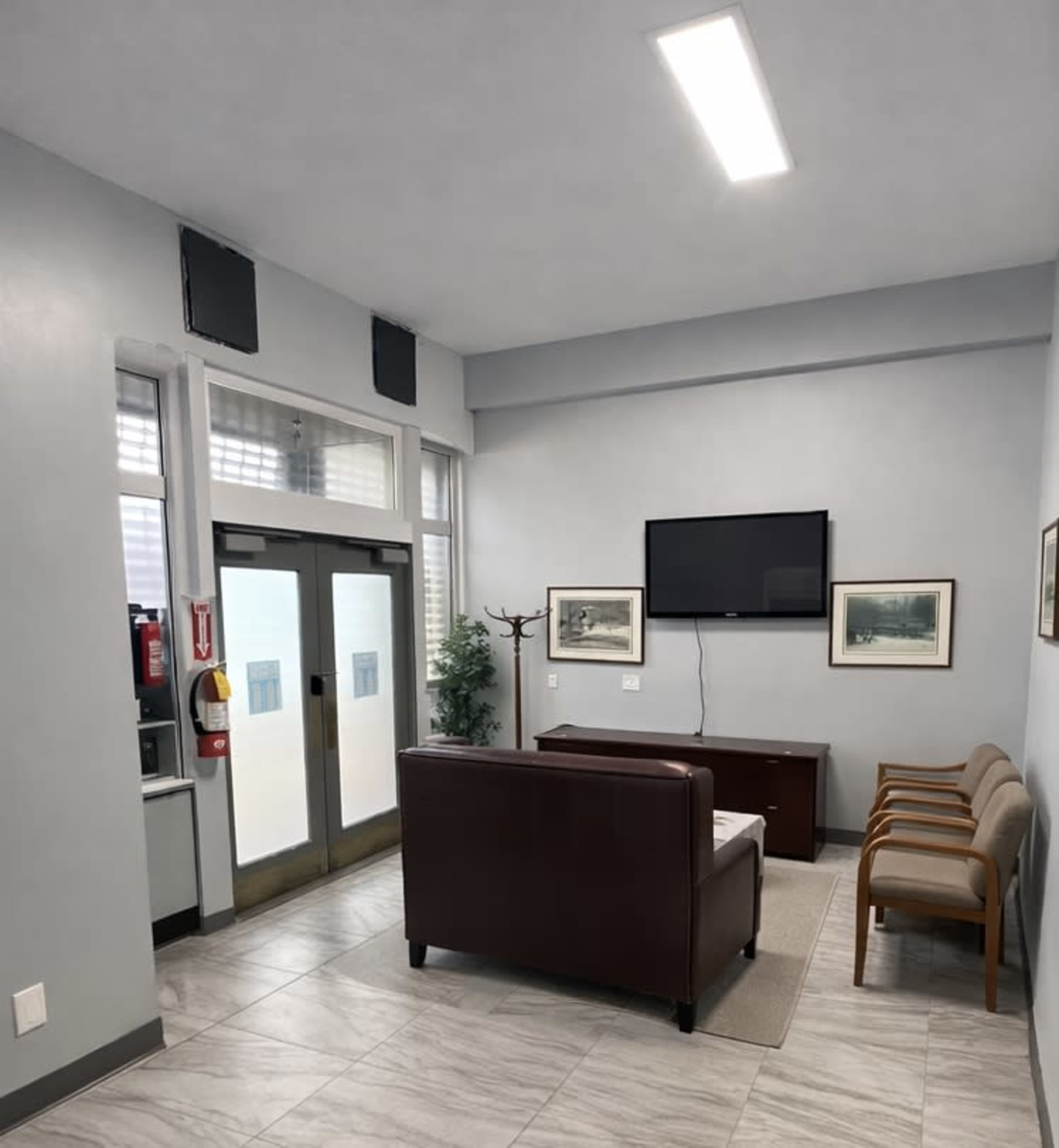 The image shows a waiting area with a brown couch, two light-colored chairs, and a television mounted on the wall.