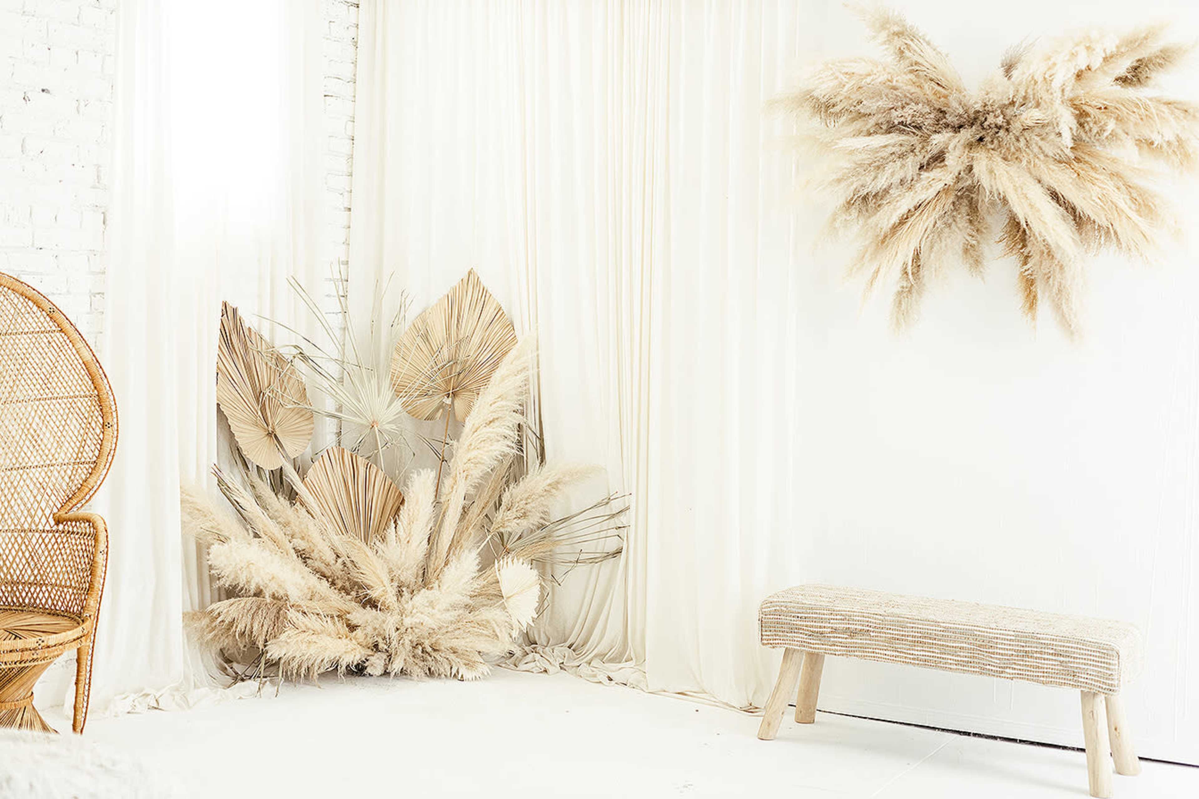 A bright, minimalist interior with large dried palm leaves arranged against a white backdrop, complemented by a woven chair and a simple bench.