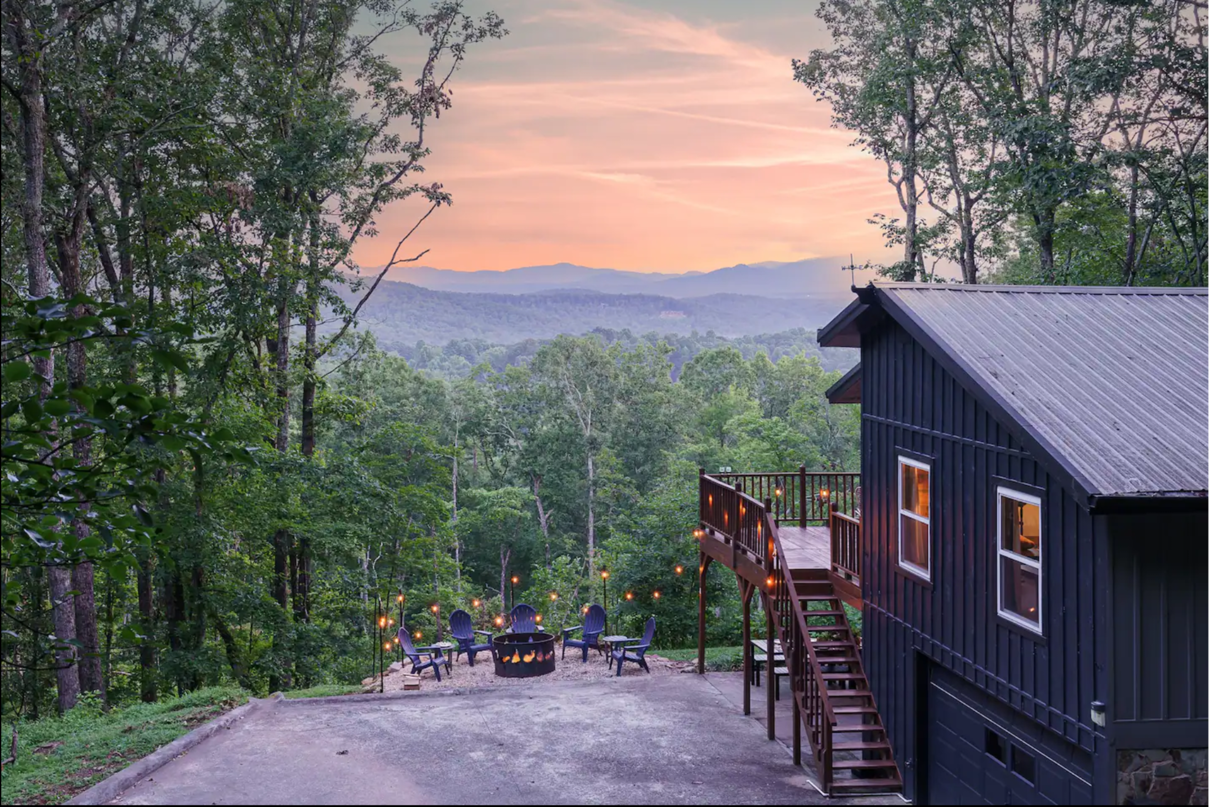 A mountain cabin overlooks a wooded valley at sunset, with a fire pit and seating area visible in the foreground.