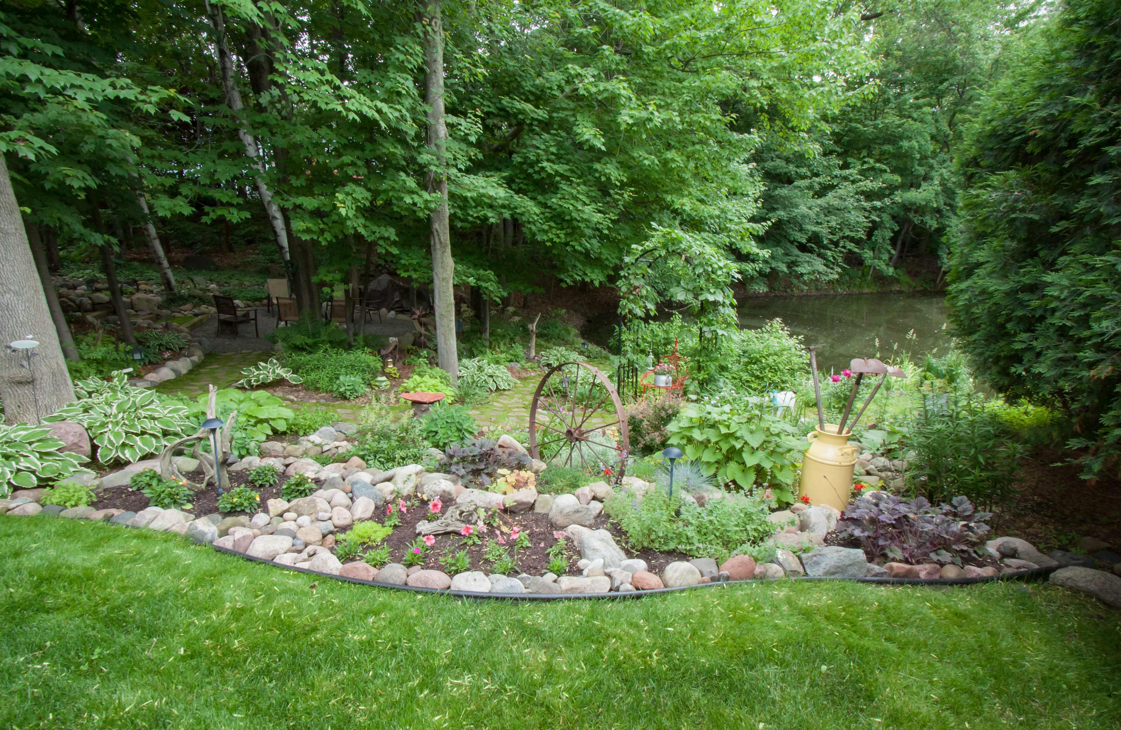 A landscaped garden features a winding path lined with stones, various plants, an old wheel, and a few colorful decorative items beside a pond.