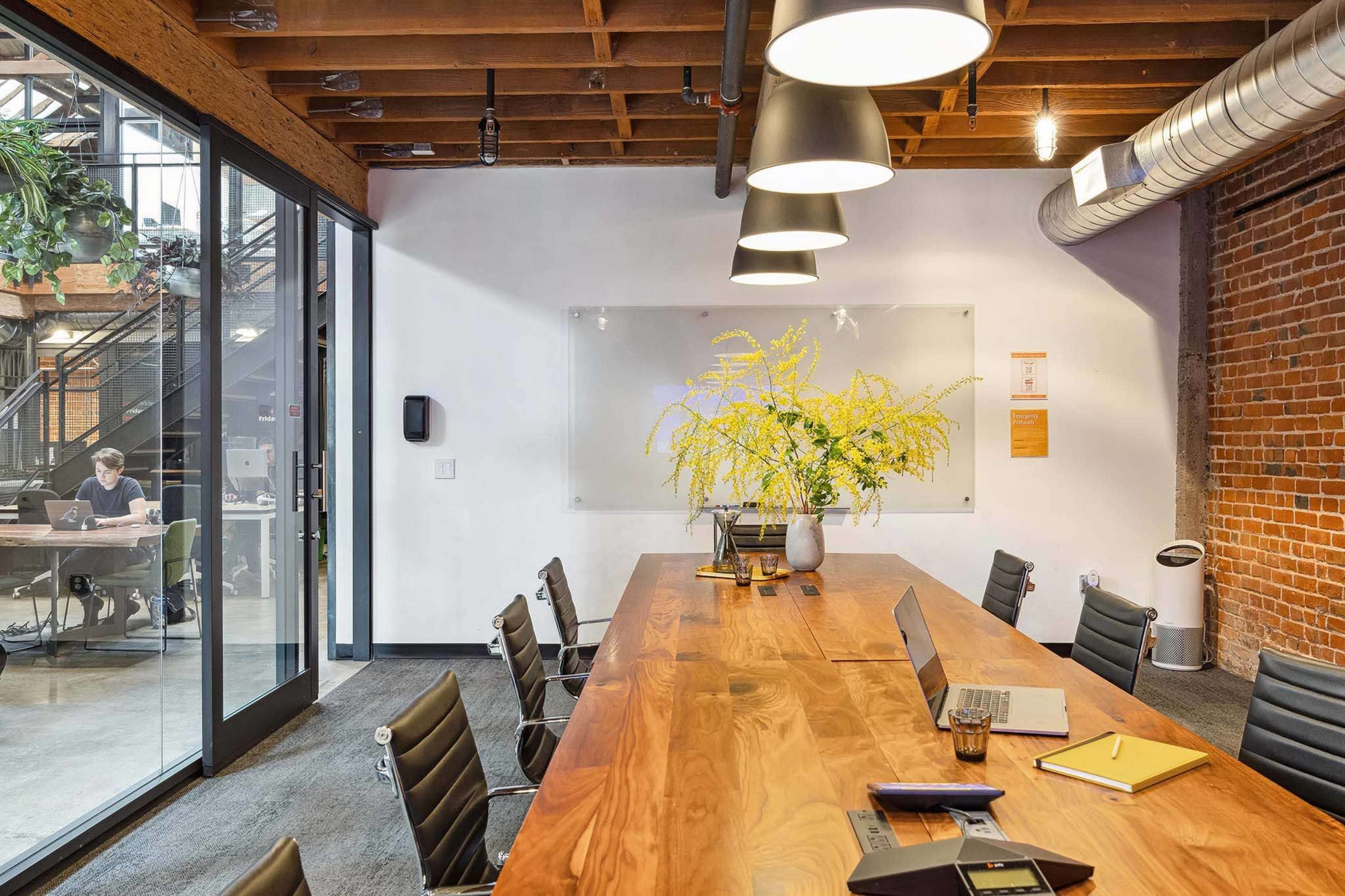 A modern conference room features a long wooden table with chairs, a large glass wall, and a floral centerpiece, while visible through the glass is a coworking space with a person working at a desk.