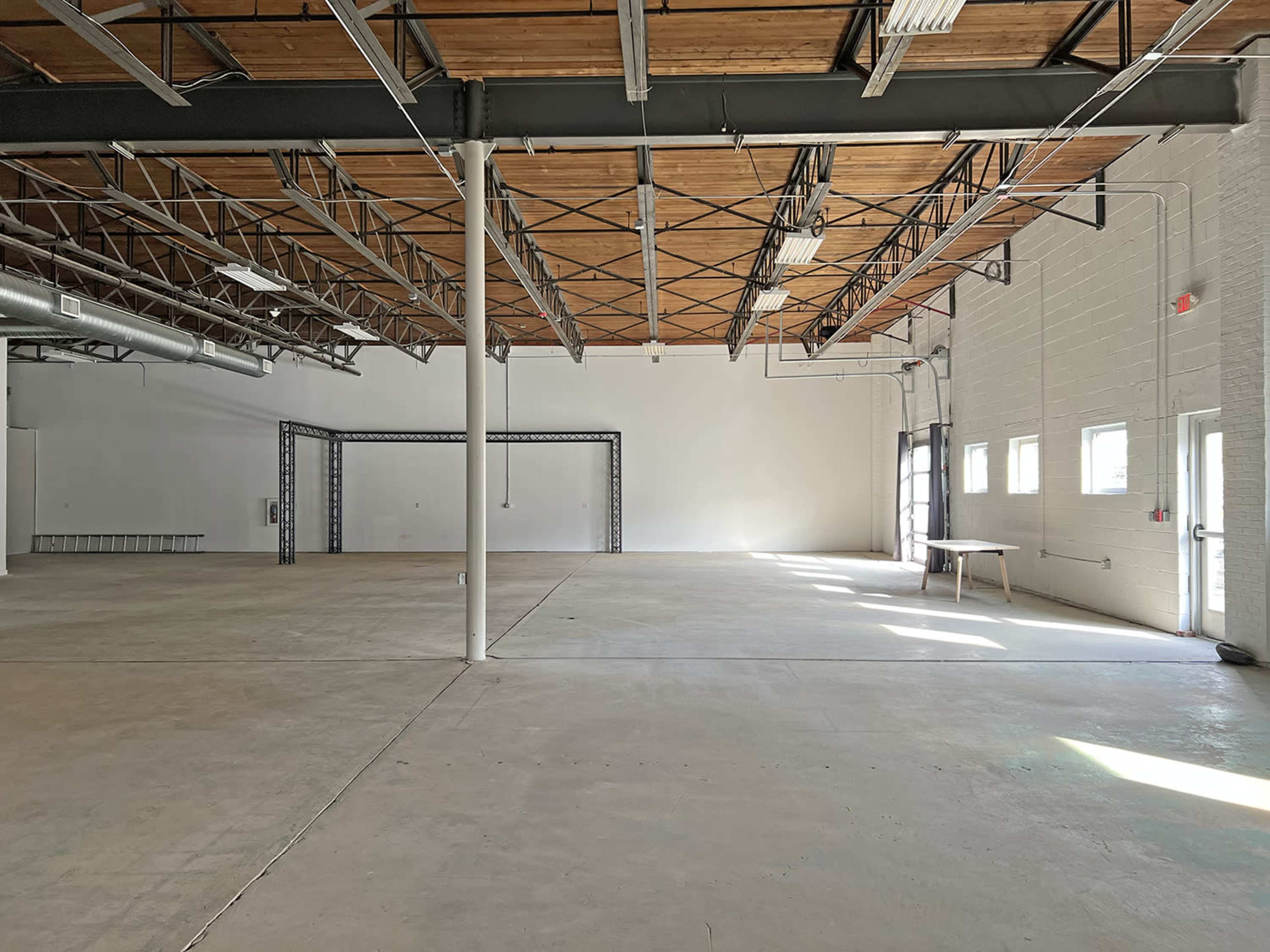 An empty industrial space with exposed beams, concrete flooring, and windows allowing natural light.