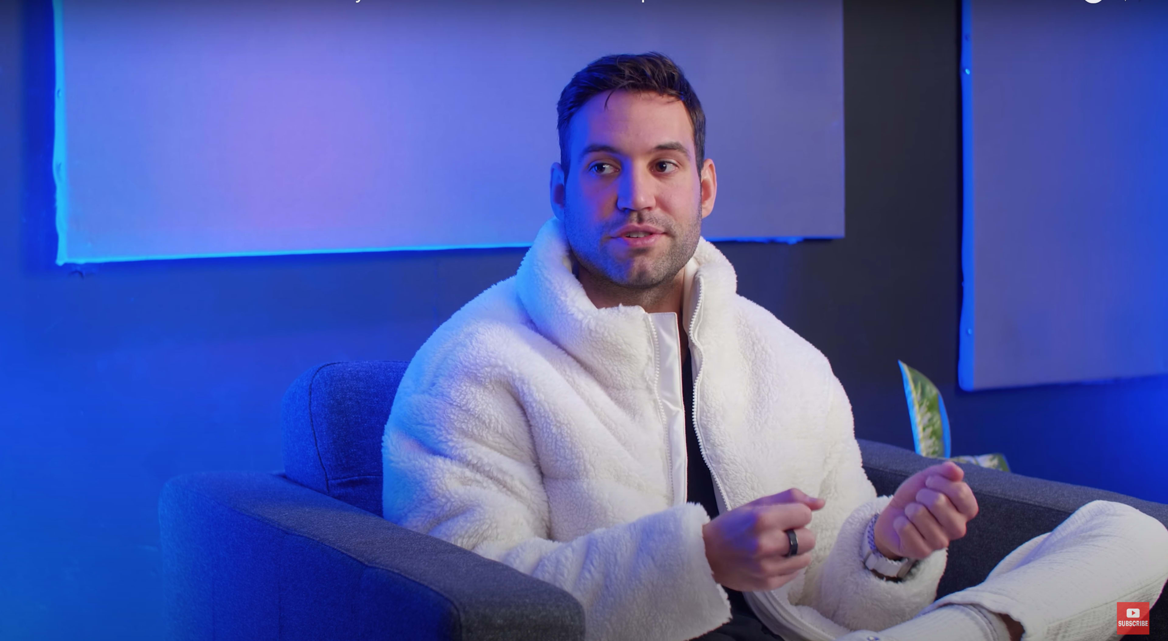 A man wearing a white jacket sits on a gray couch while speaking in a room illuminated by blue light.