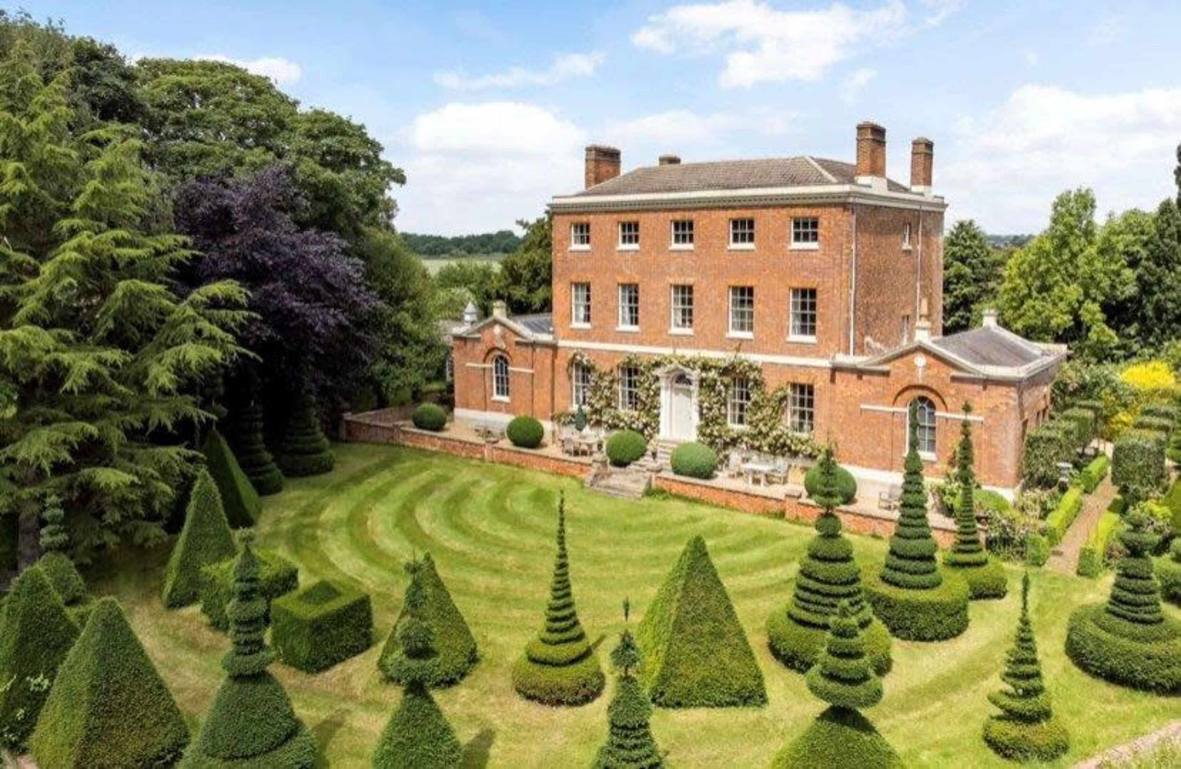 A large red brick mansion is surrounded by meticulously trimmed topiary in a landscaped garden.
