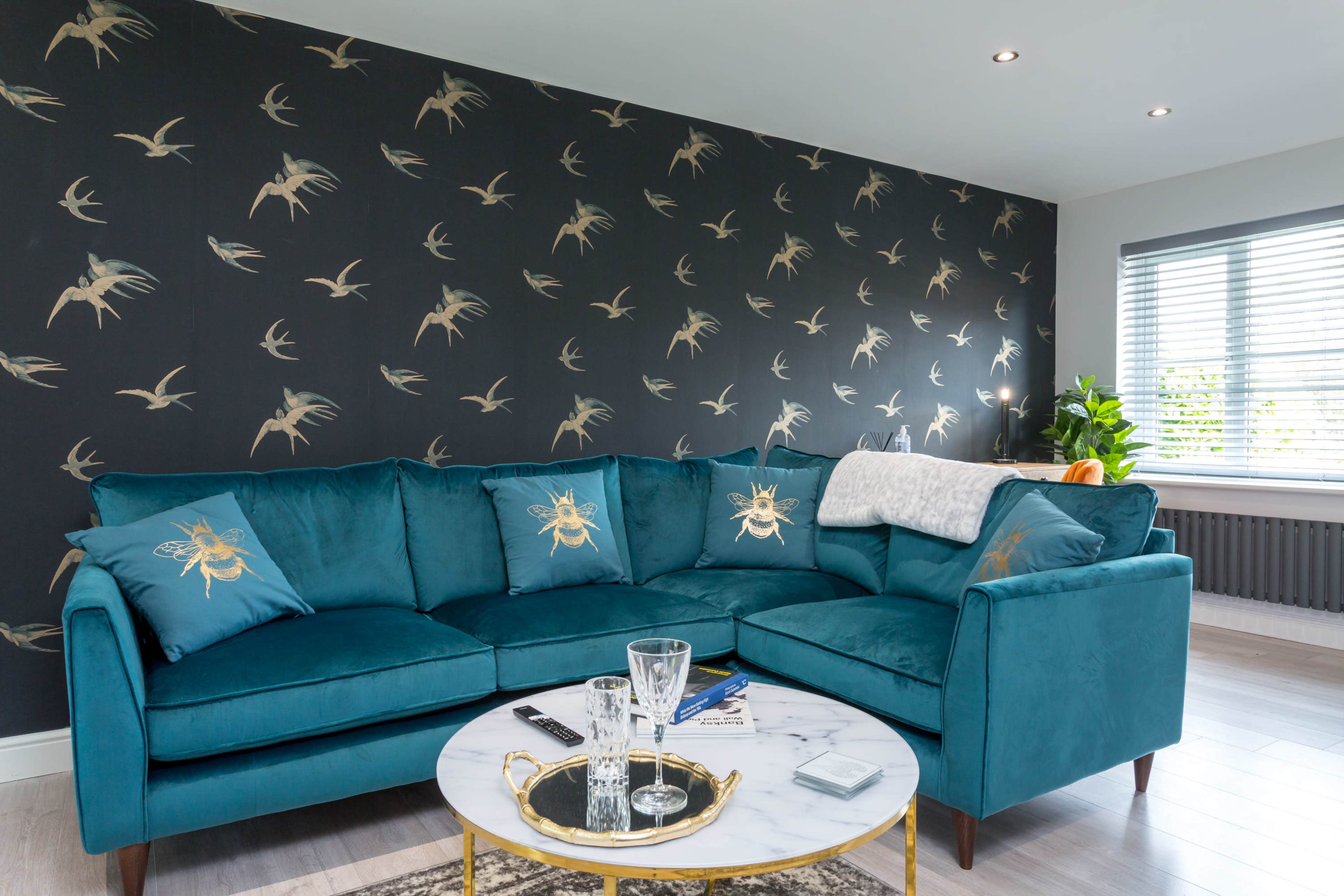 A teal sectional sofa with decorative cushions is positioned in a living room featuring a patterned wallpaper of birds and a marble coffee table.