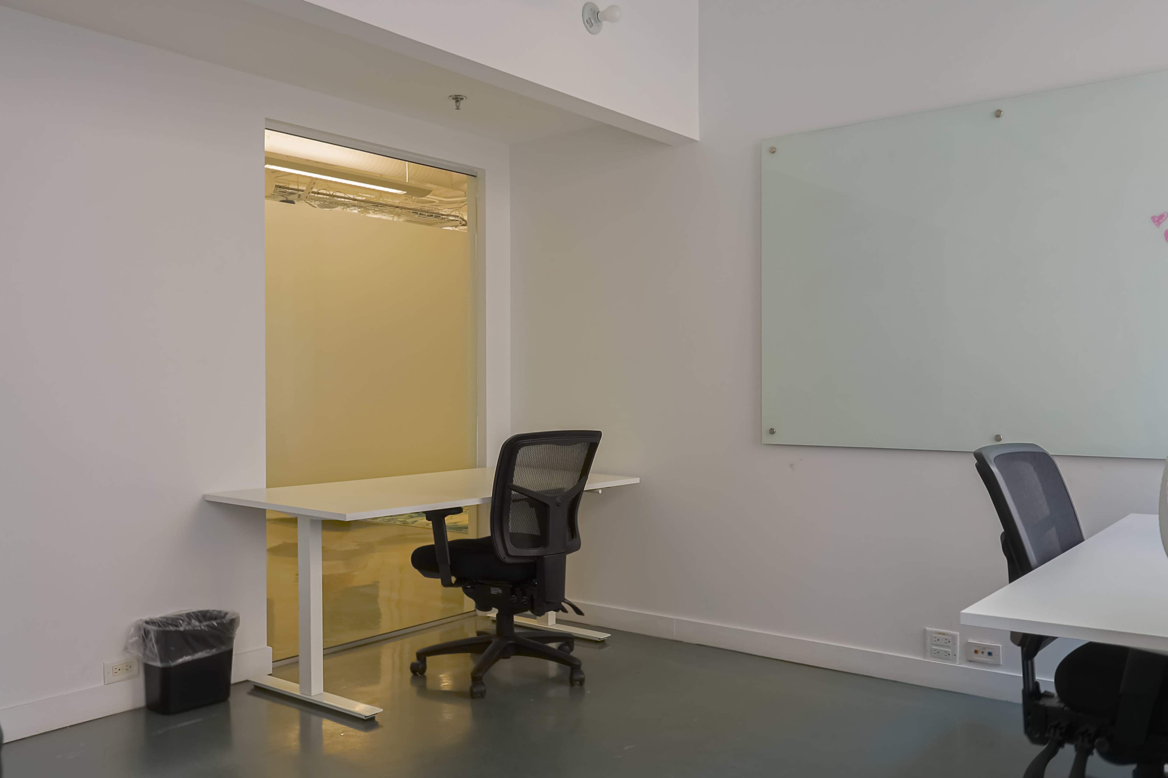 A modern office space features a clean, minimalistic design with a white desk and black ergonomic chair, along with a glass panel that leads to a separate room.