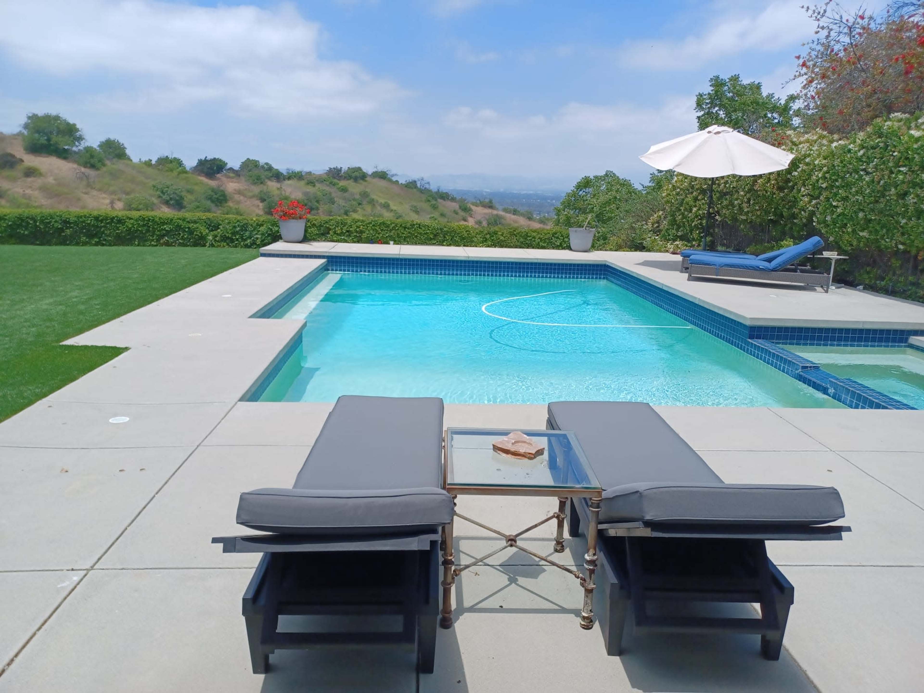 A swimming pool with chaise lounges and a side table is set against a backdrop of green hills under a partly cloudy sky.