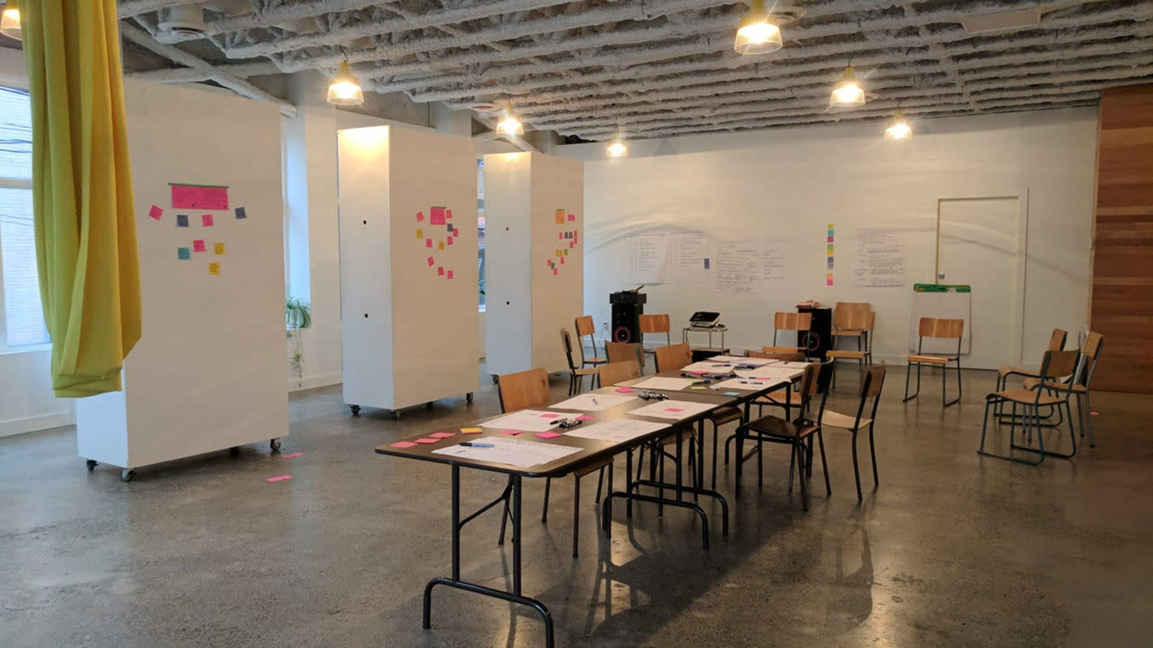 A spacious room features a long table with papers and sticky notes, surrounded by several chairs and movable whiteboards displaying colorful notes.