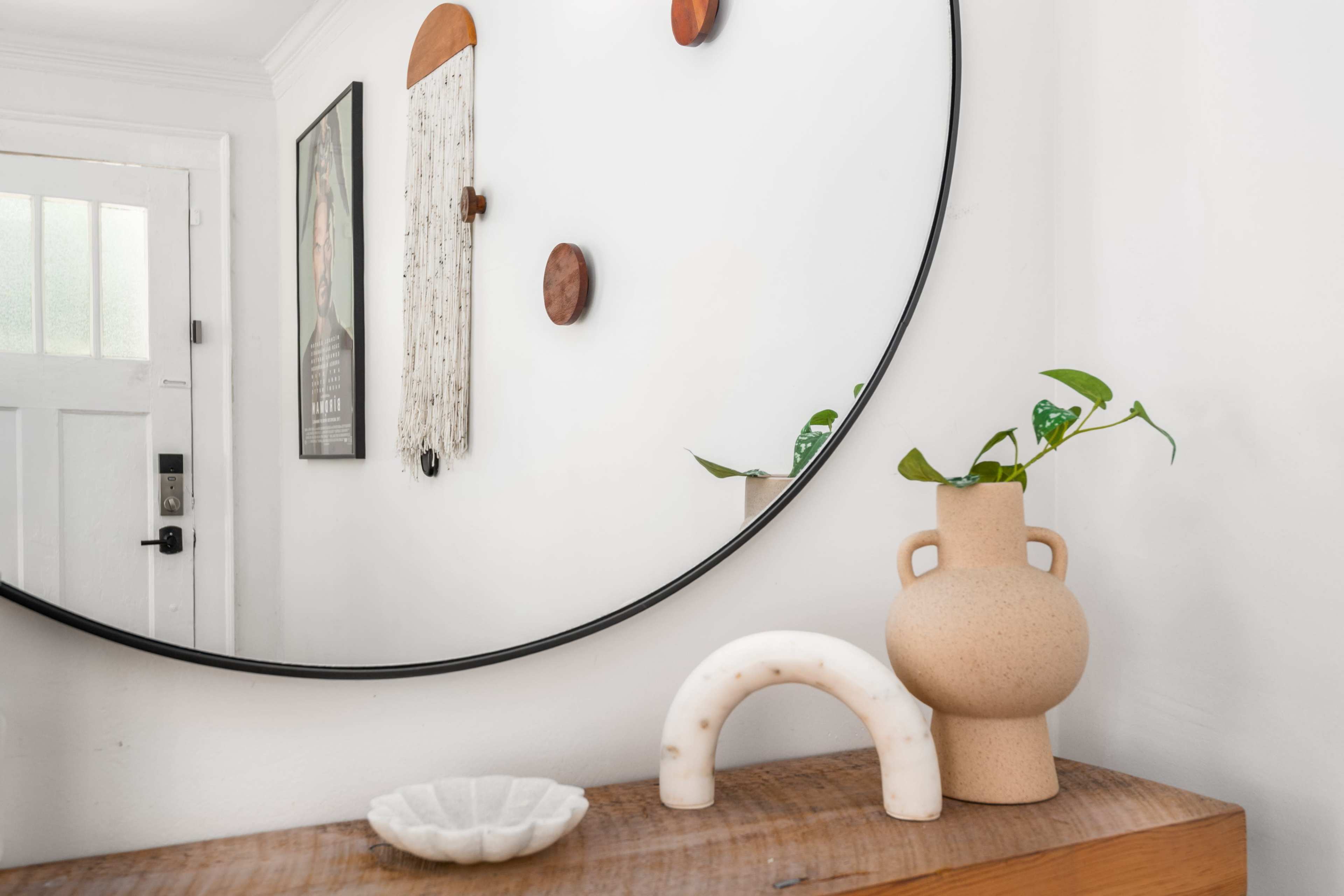 The image shows a modern entryway with a round mirror reflecting a wall with decorative items, a wooden console table, a ceramic vase, and a small plant.