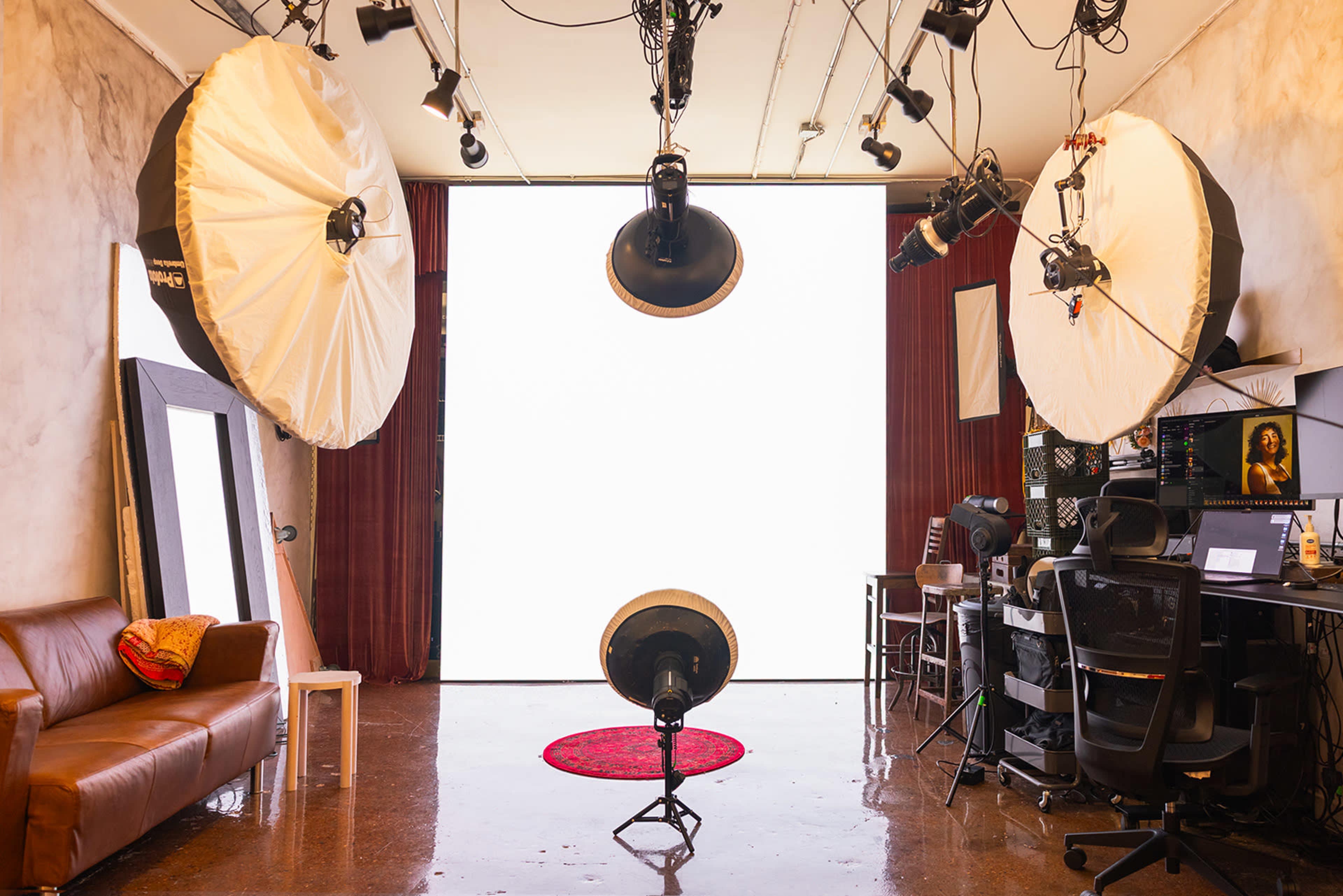 The image shows a photography studio with two large softbox lights, a round red rug on the floor, and a white backdrop, along with various lighting equipment and a computer setup.