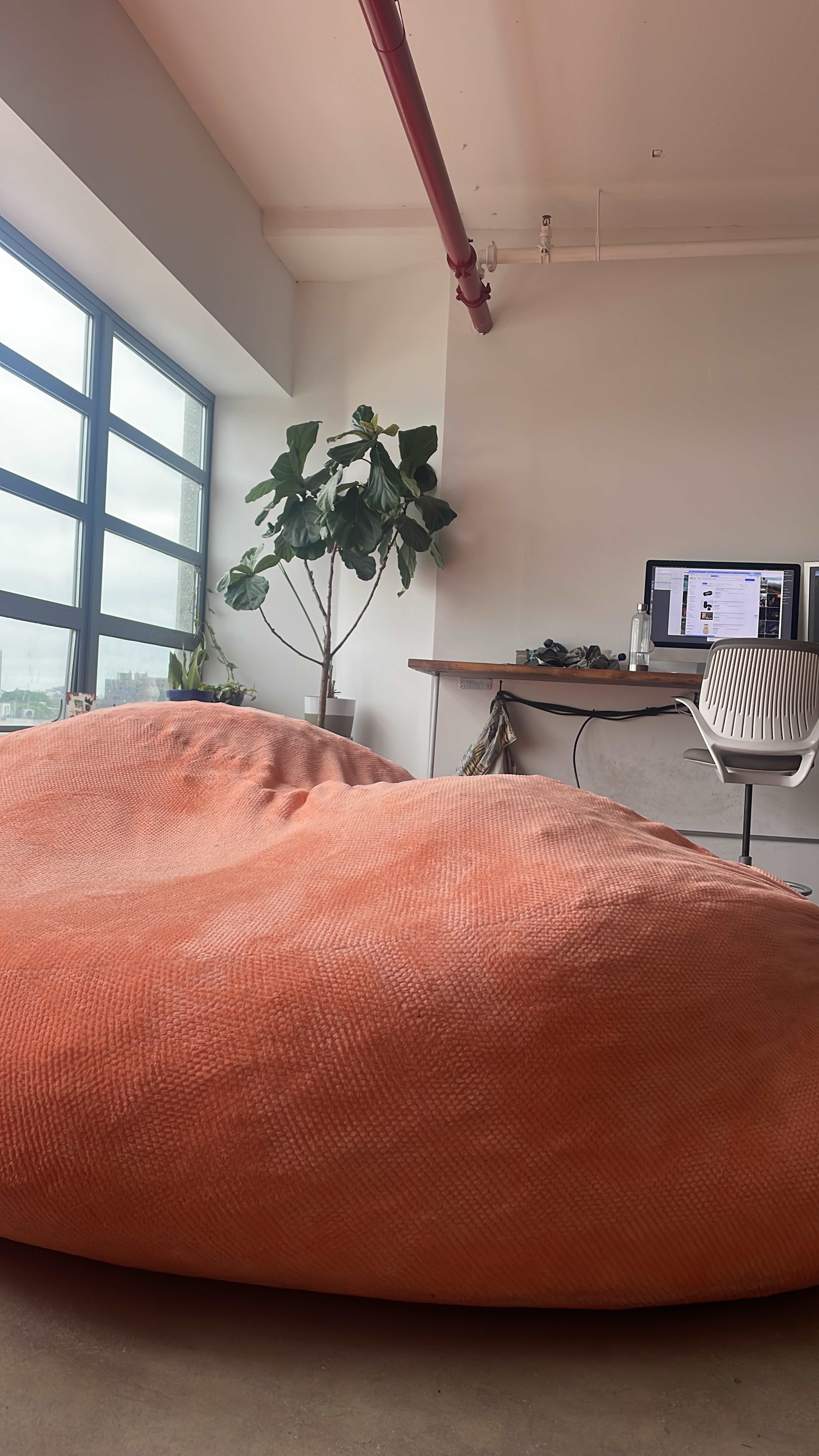 A large orange bean bag sits in a modern office space with a desk, computer, and a potted plant near a window.