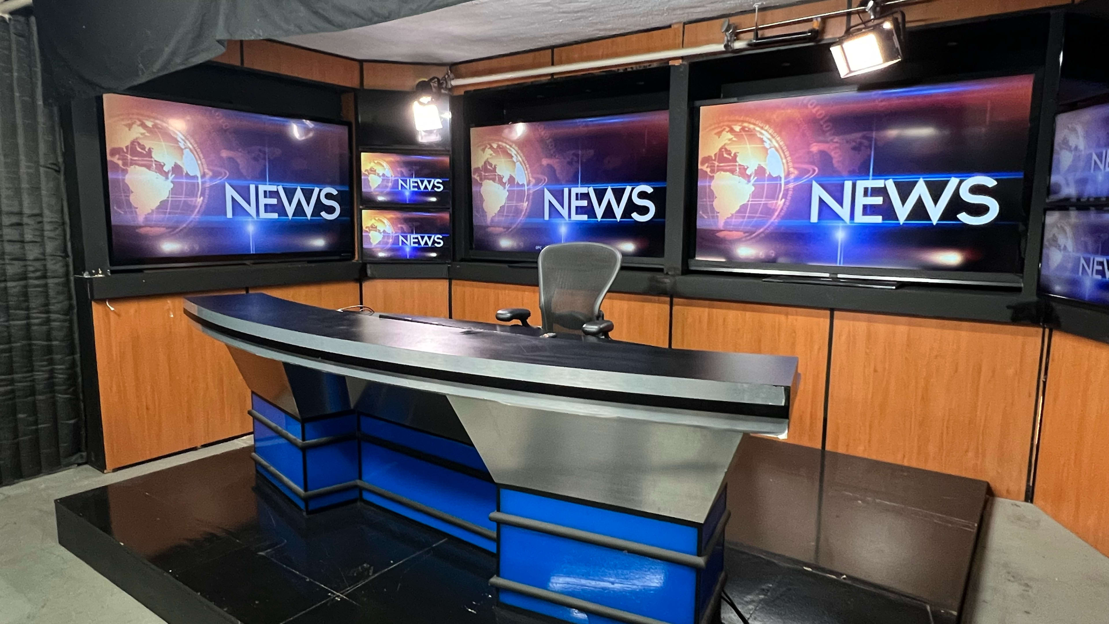 The image shows a television news studio with multiple screens displaying the word "NEWS" and a modern desk with a chair.