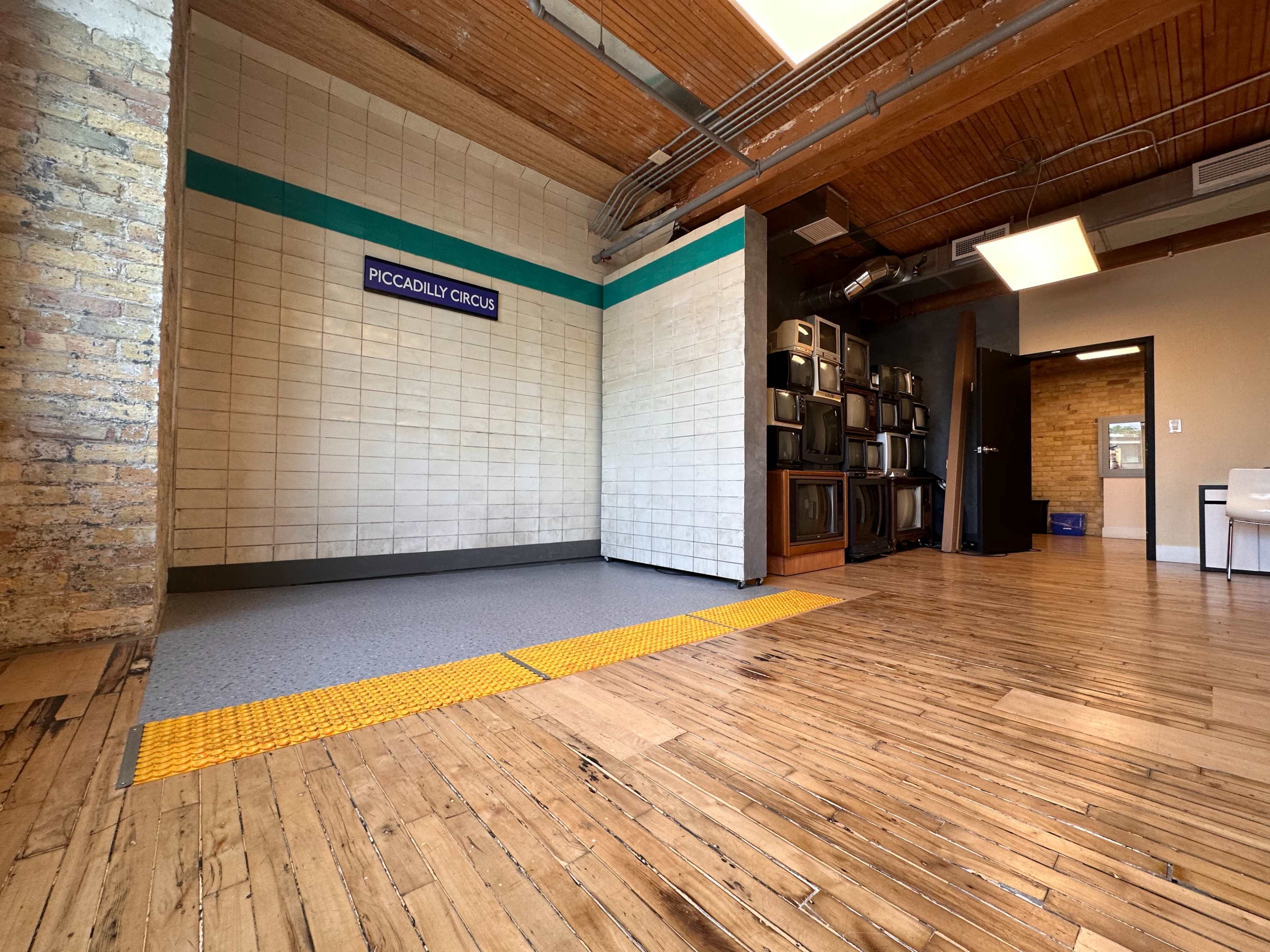 The image shows a spacious interior with wooden flooring, exposed brick walls, and a corner featuring a sign that reads "PICCADILLY CIRCUS," along with a row of vintage televisions.