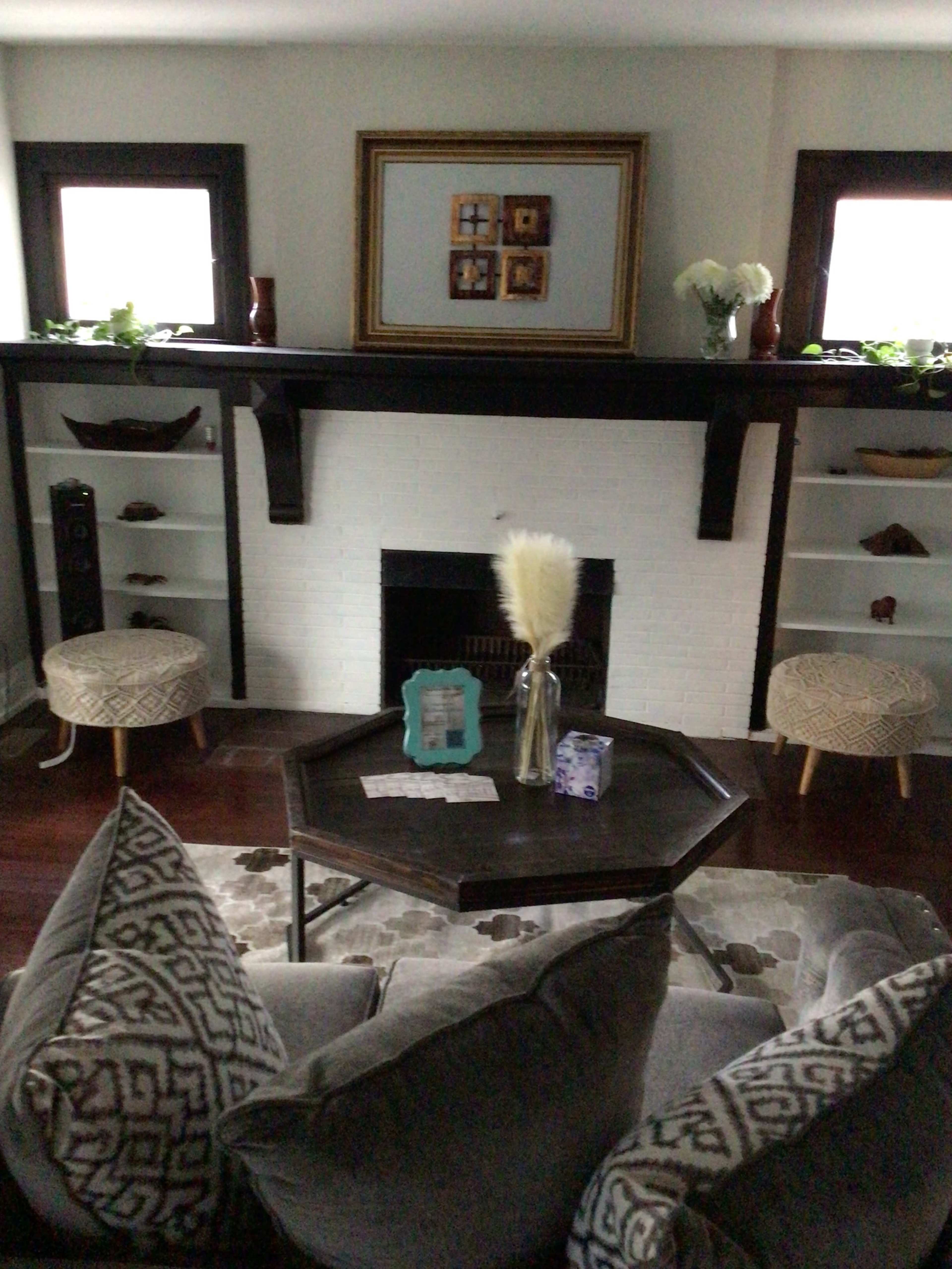 A cozy living room features a central coffee table, two patterned stools, and a mantel with framed photos above a white brick fireplace.