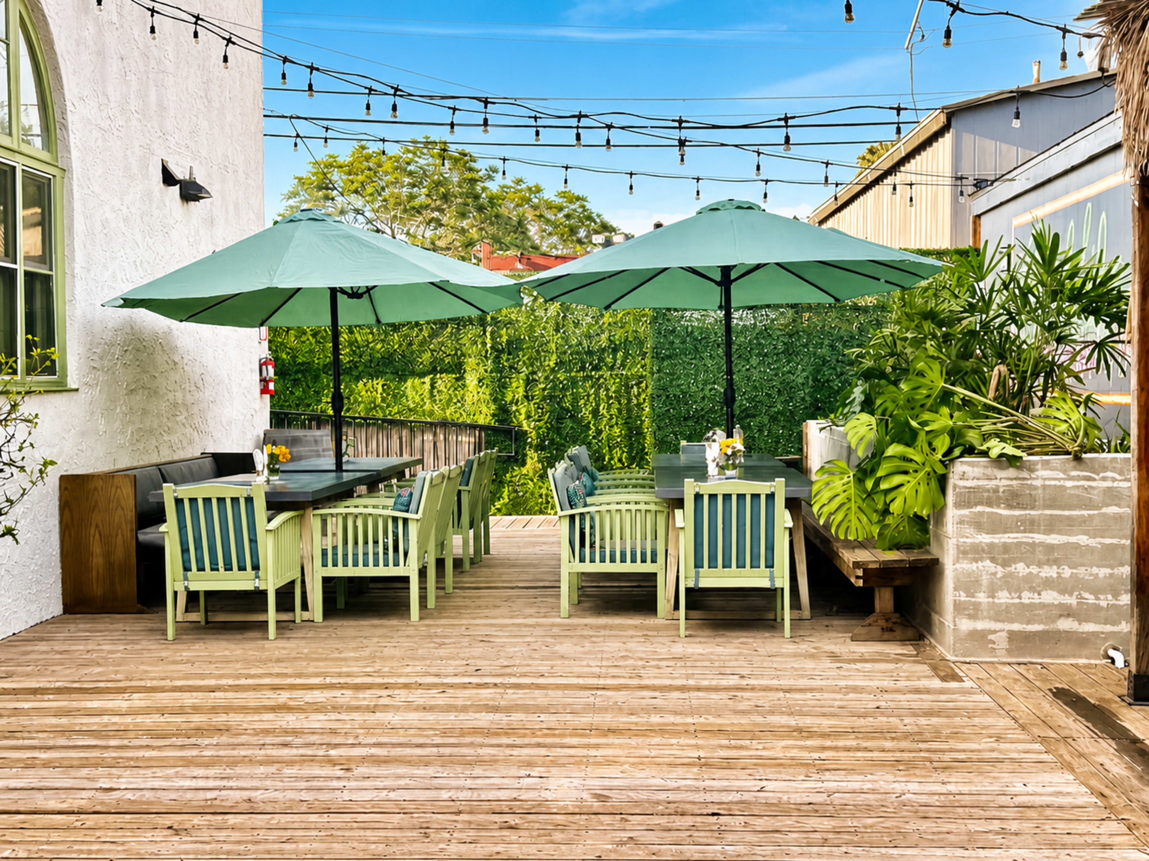 The image shows an outdoor dining area featuring several wooden tables and green umbrellas, surrounded by greenery and wooden decking.