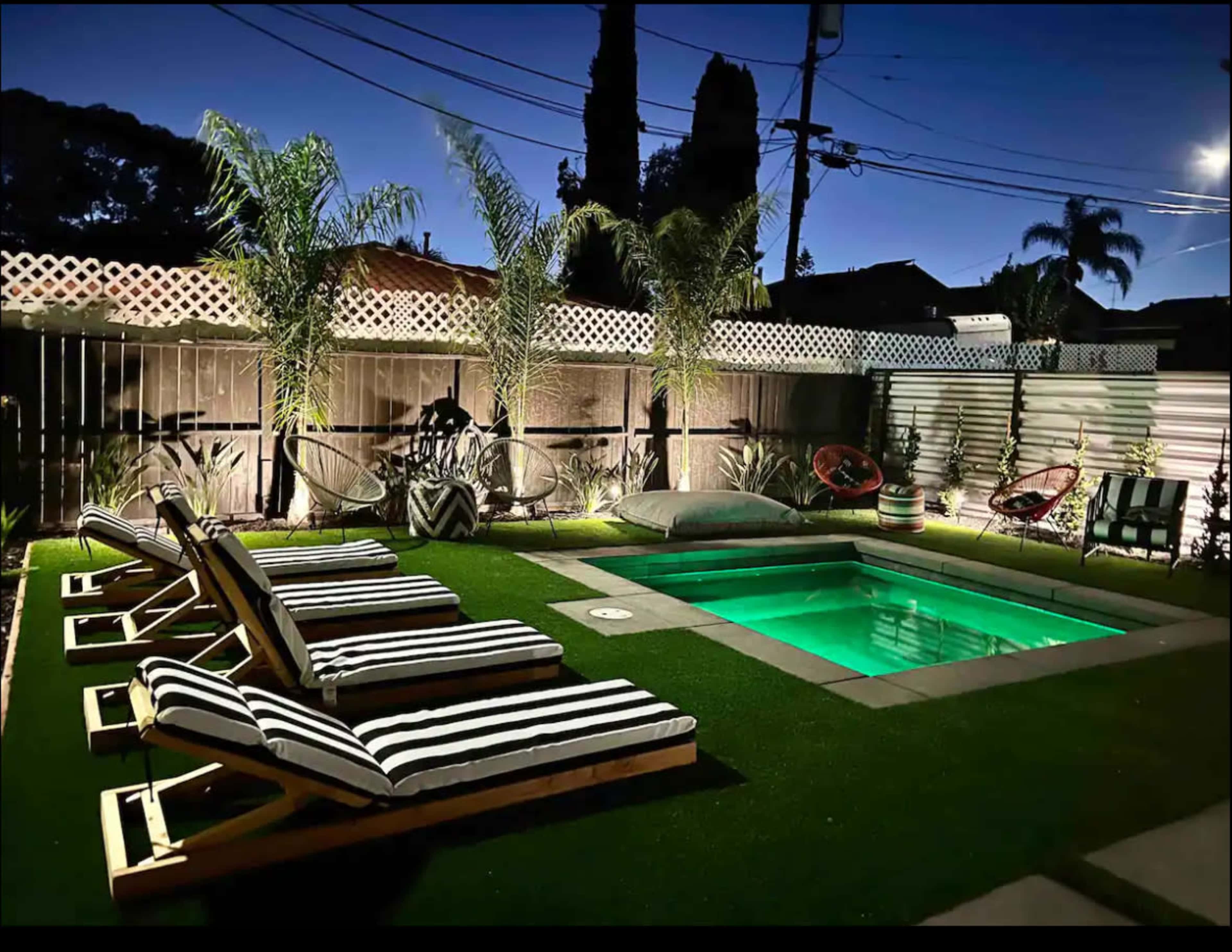 The image shows a modern backyard with a small illuminated pool surrounded by striped lounge chairs and palm trees under a twilight sky.