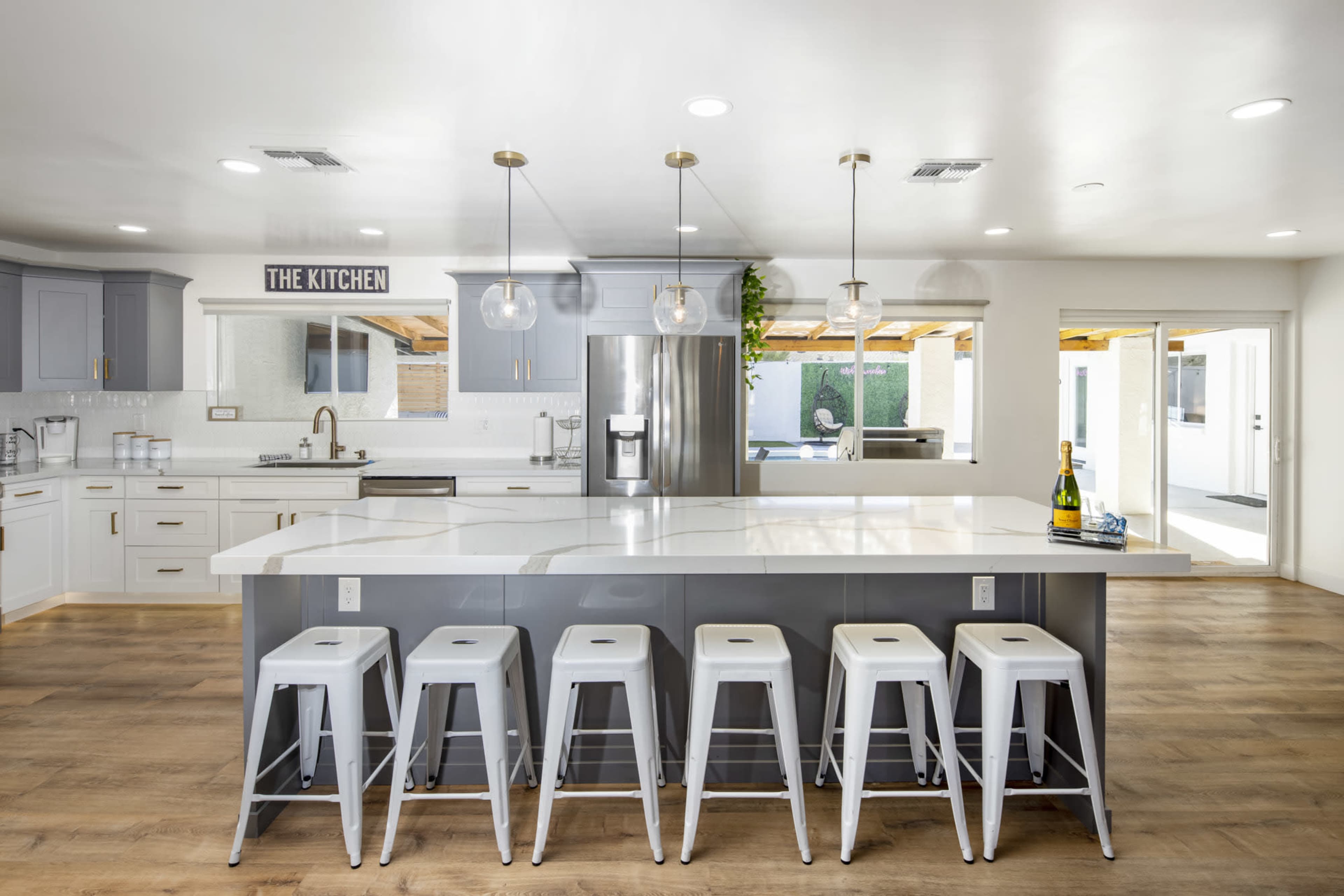 A modern kitchen features a large island with five metal stools, stainless steel appliances, and bright, open space leading to an outdoor area.
