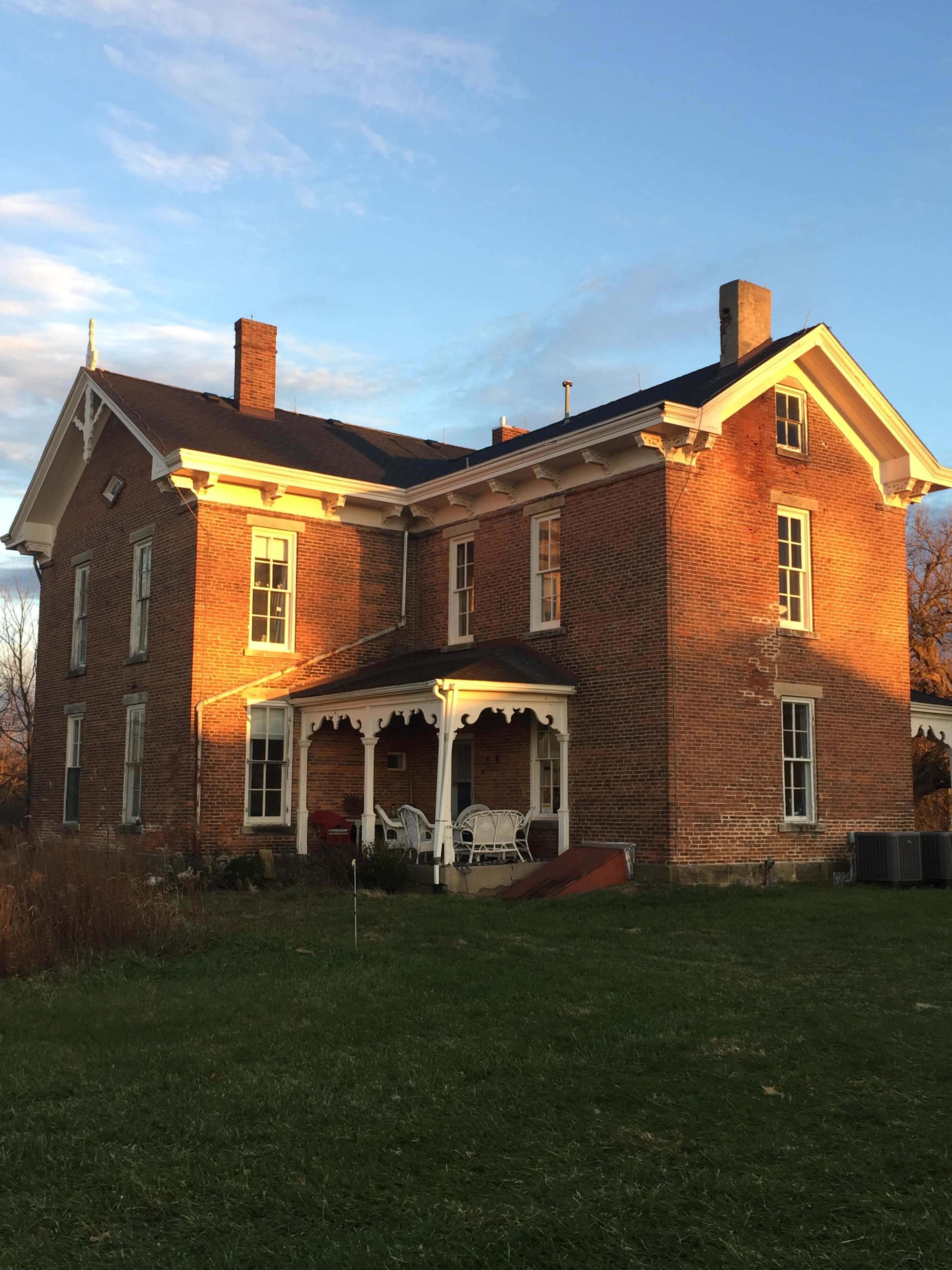 A two-story brick house with a porch and several windows, set against a backdrop of a clear sky and grassy lawn.