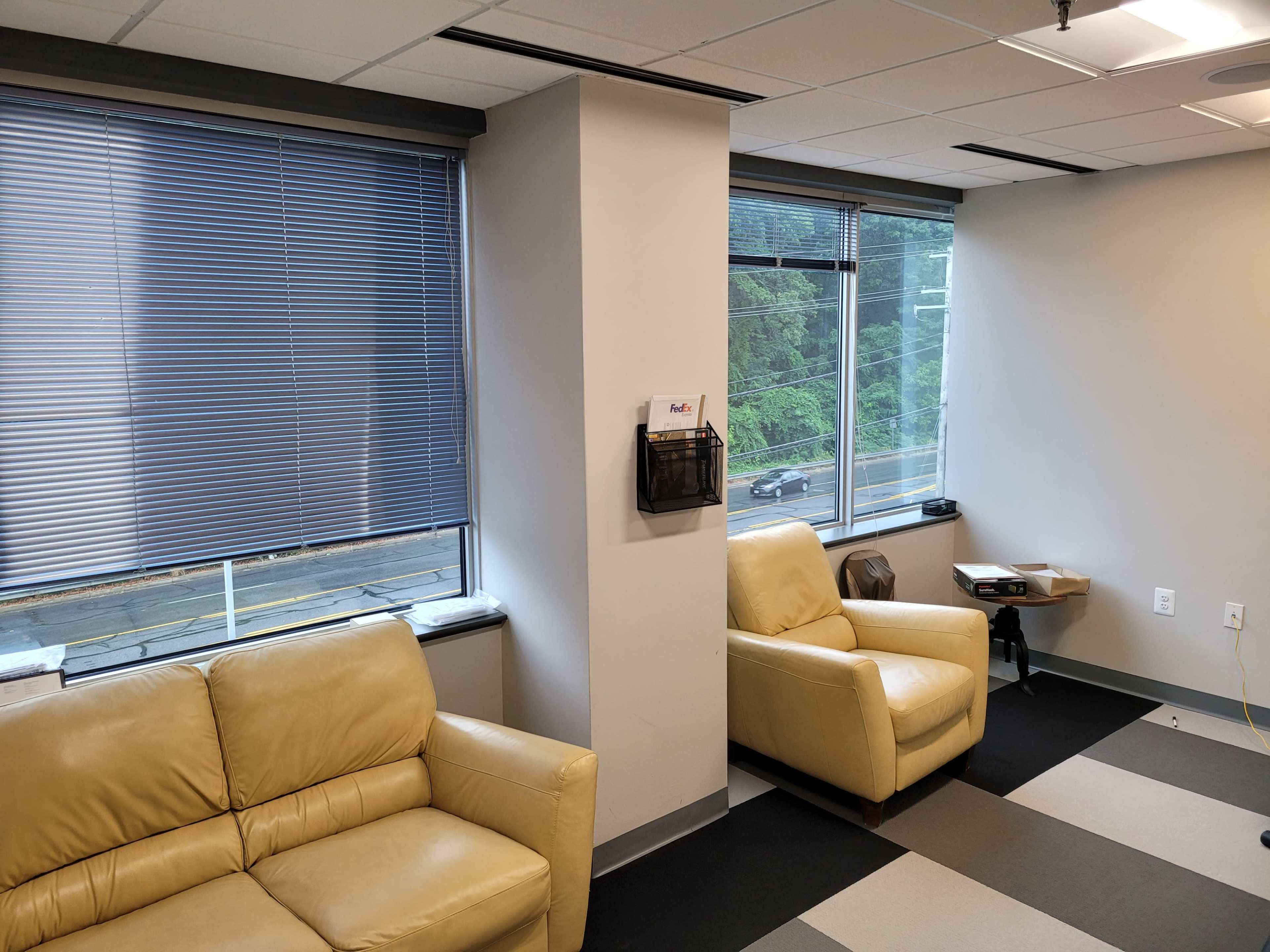 A corner of an office with two light-colored armchairs, large windows covered by blinds, and a wall-mounted pamphlet holder.