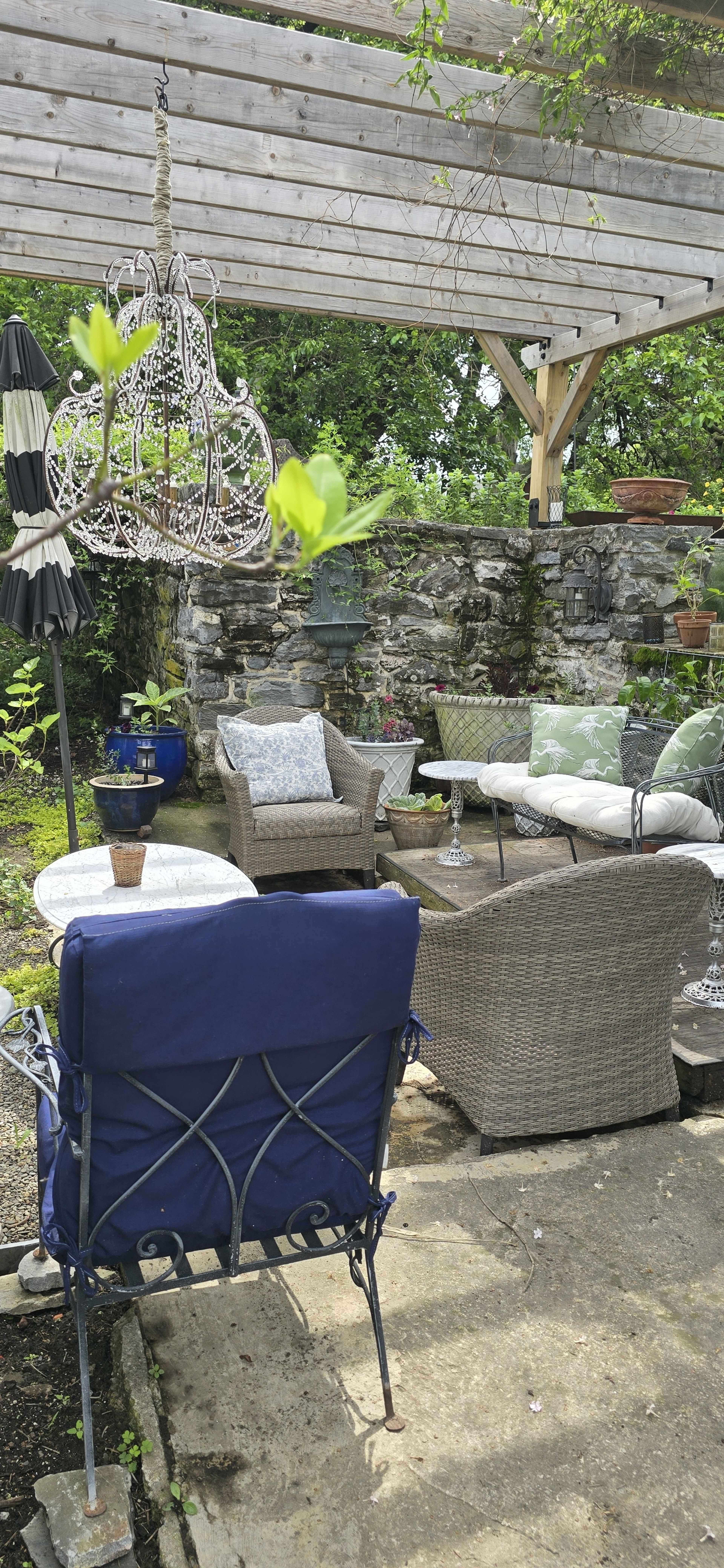 A shaded outdoor seating area features woven furniture, a stone wall backdrop, and potted plants.