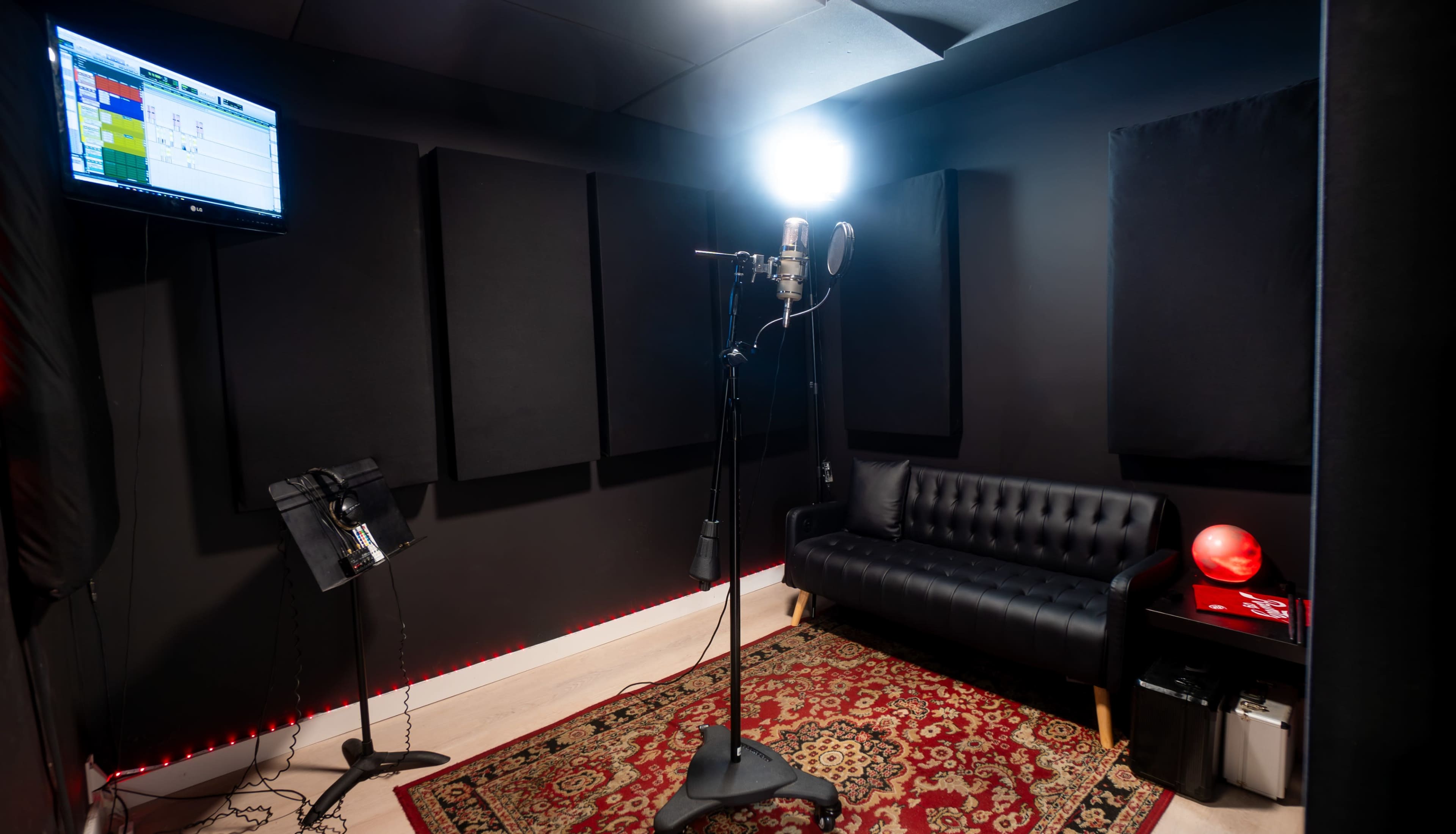 The image shows a soundproof recording studio with a microphone on a stand, a black sofa, a red lamp, and acoustic panels on the walls.