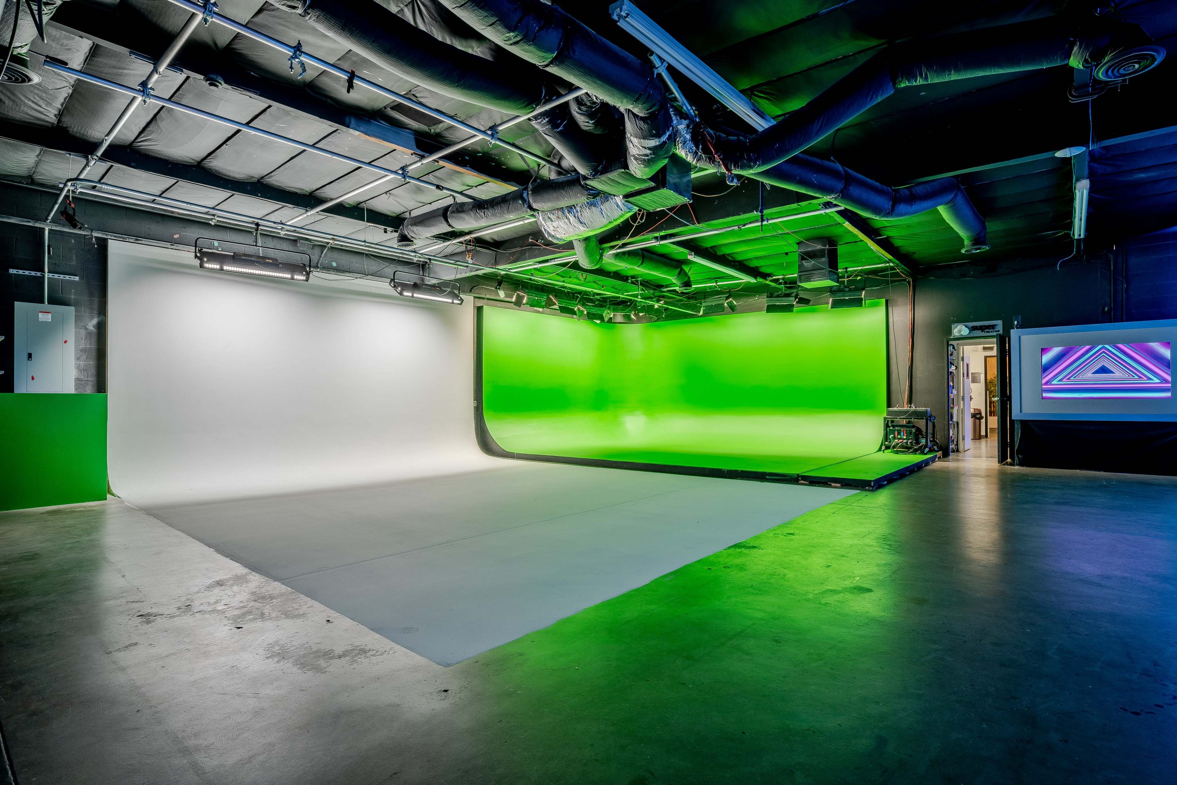 A photography studio featuring a white backdrop on one side and a green screen on the other, with pipes and lights visible above.