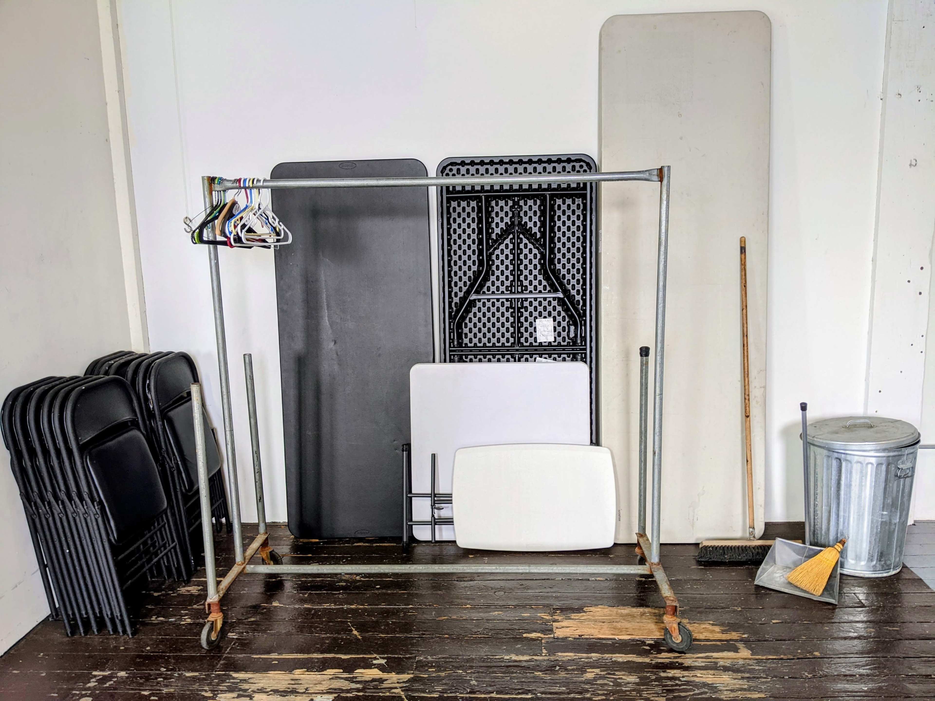 A clothing rack with hangers stands among stacked folding tables, a broom, and a trash can in a sparsely furnished room.