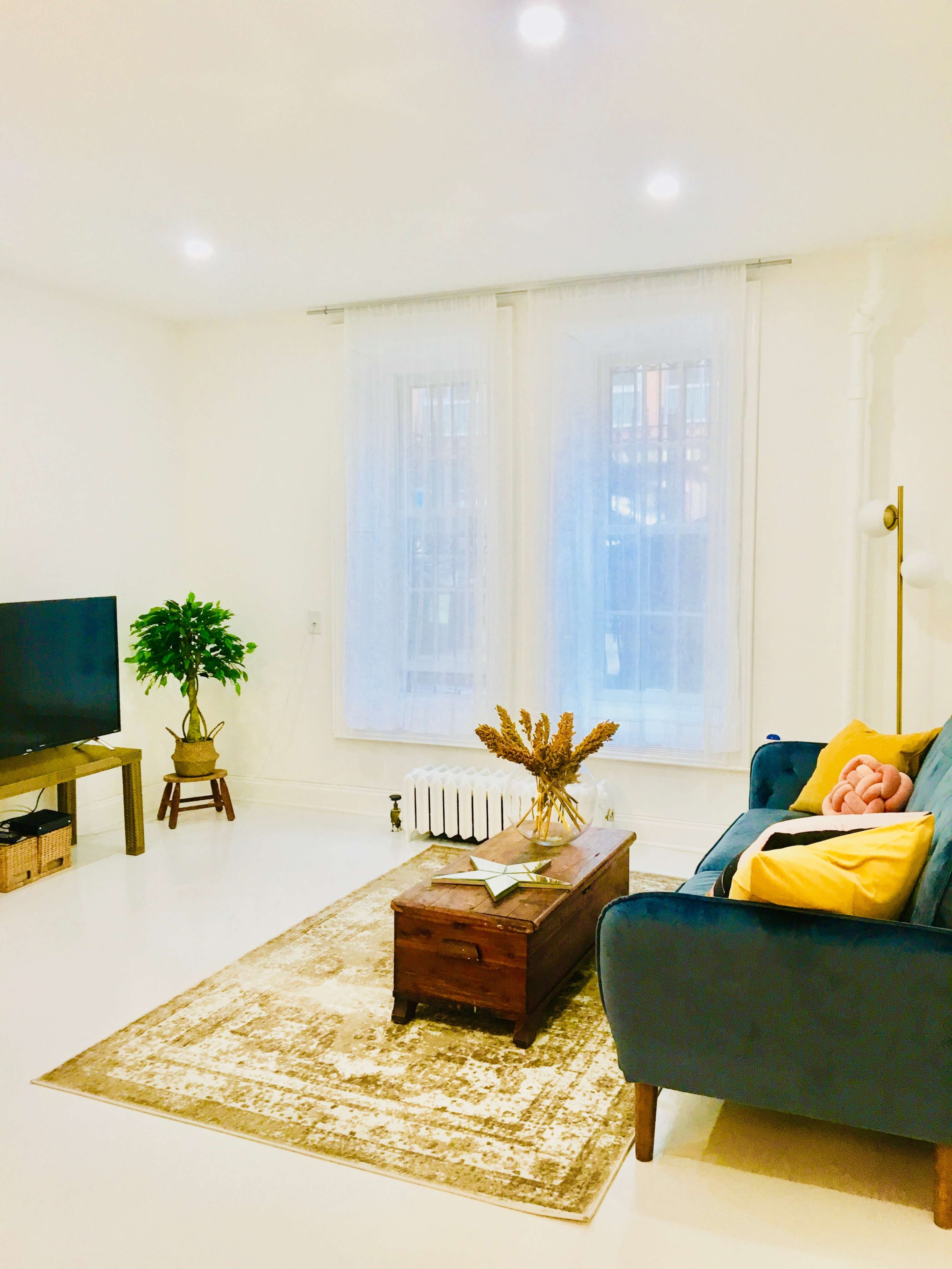 The image shows a bright living room featuring a blue sofa, a wooden coffee table with decorative items, a television on a stand, and large windows covered with sheer curtains.