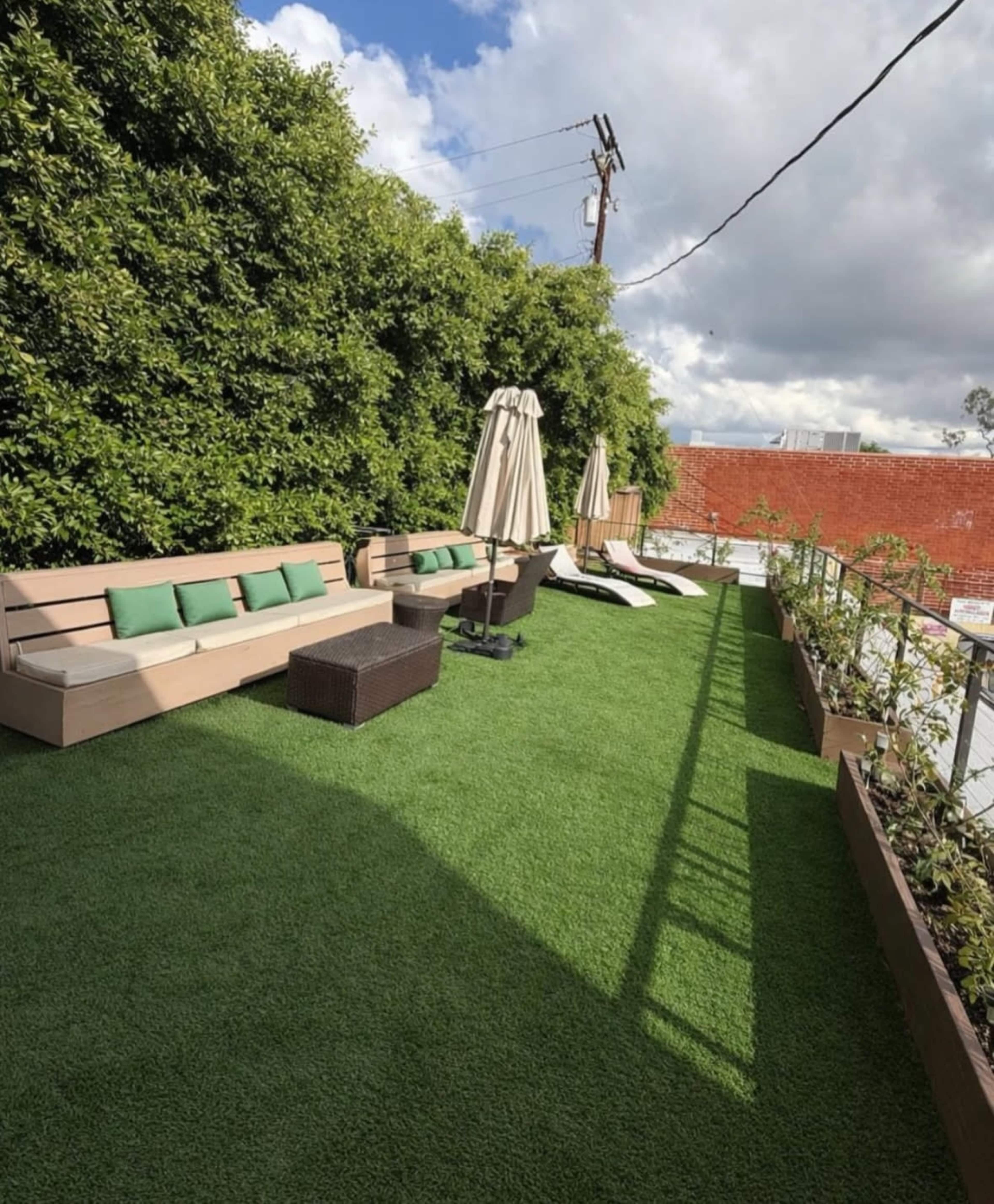 The image shows a landscaped outdoor space featuring a green artificial lawn, a sectional sofa with green cushions, a patio umbrella, a coffee table, and two lounge chairs against a backdrop of a wall and plants.