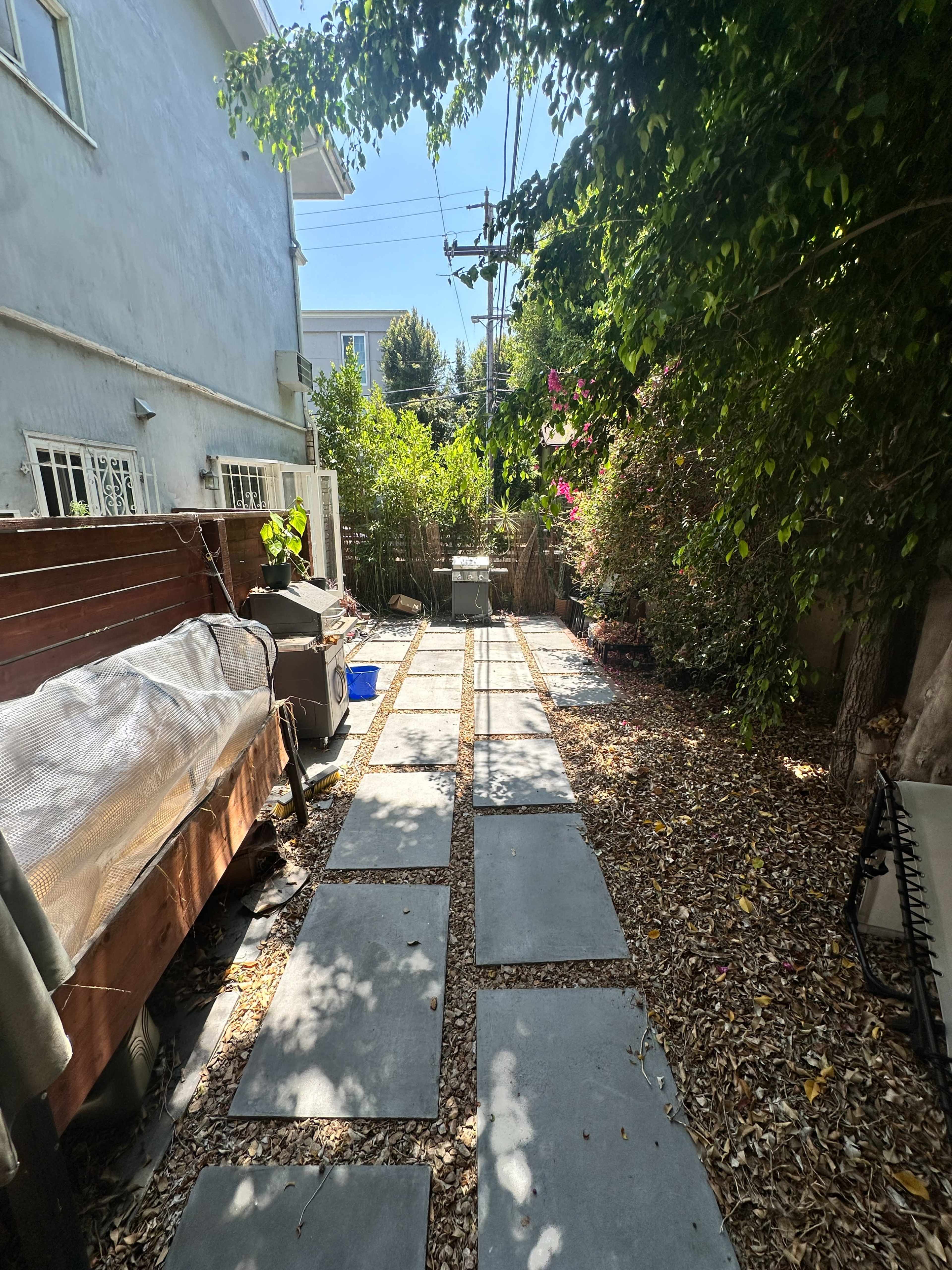 The image shows a narrow backyard pathway lined with stone slabs, flanked by greenery and a barbecue grill on one side.