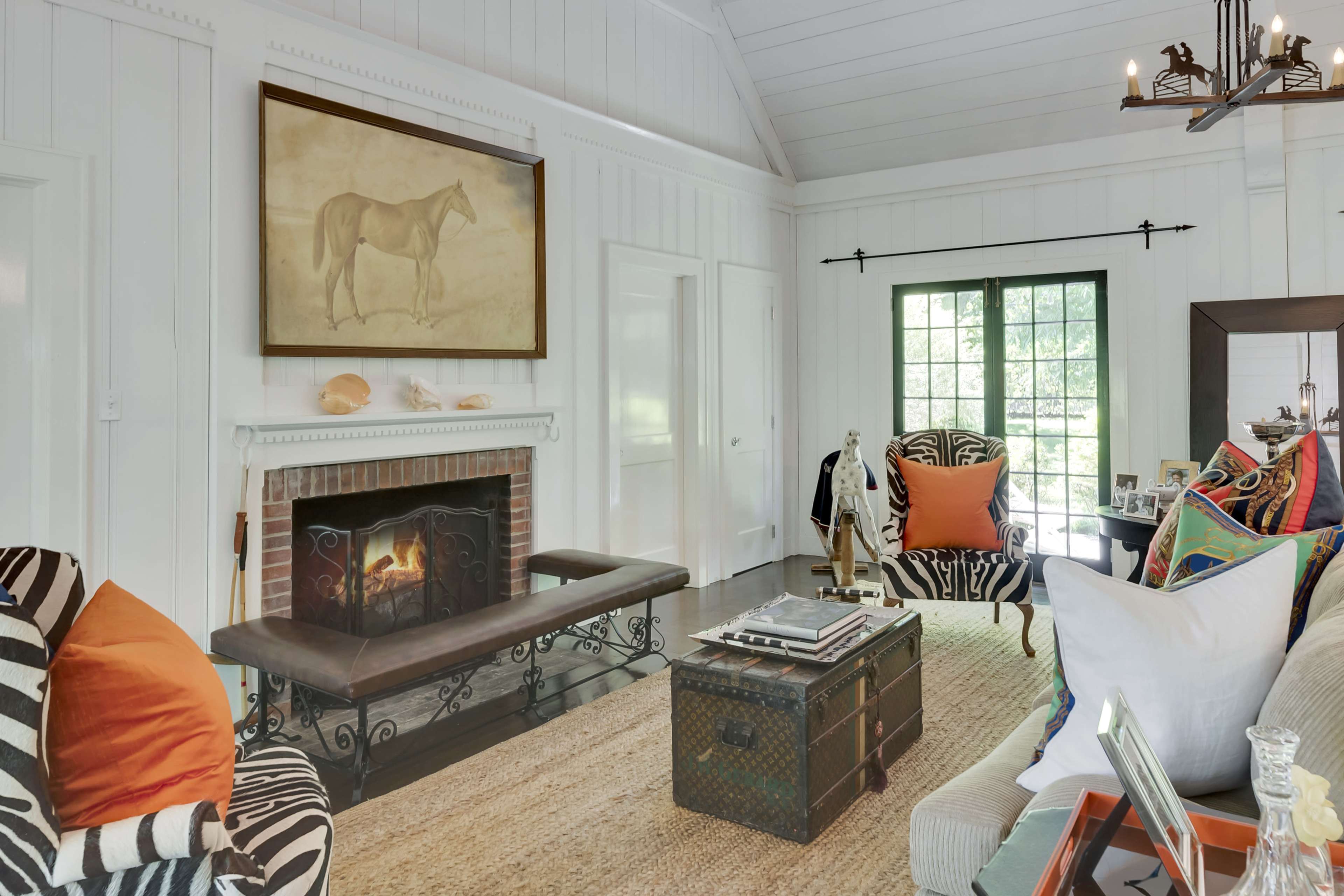The image shows a cozy living room with a fireplace, a large painting of a horse on the wall, and various patterned furniture arranged around a woven rug.