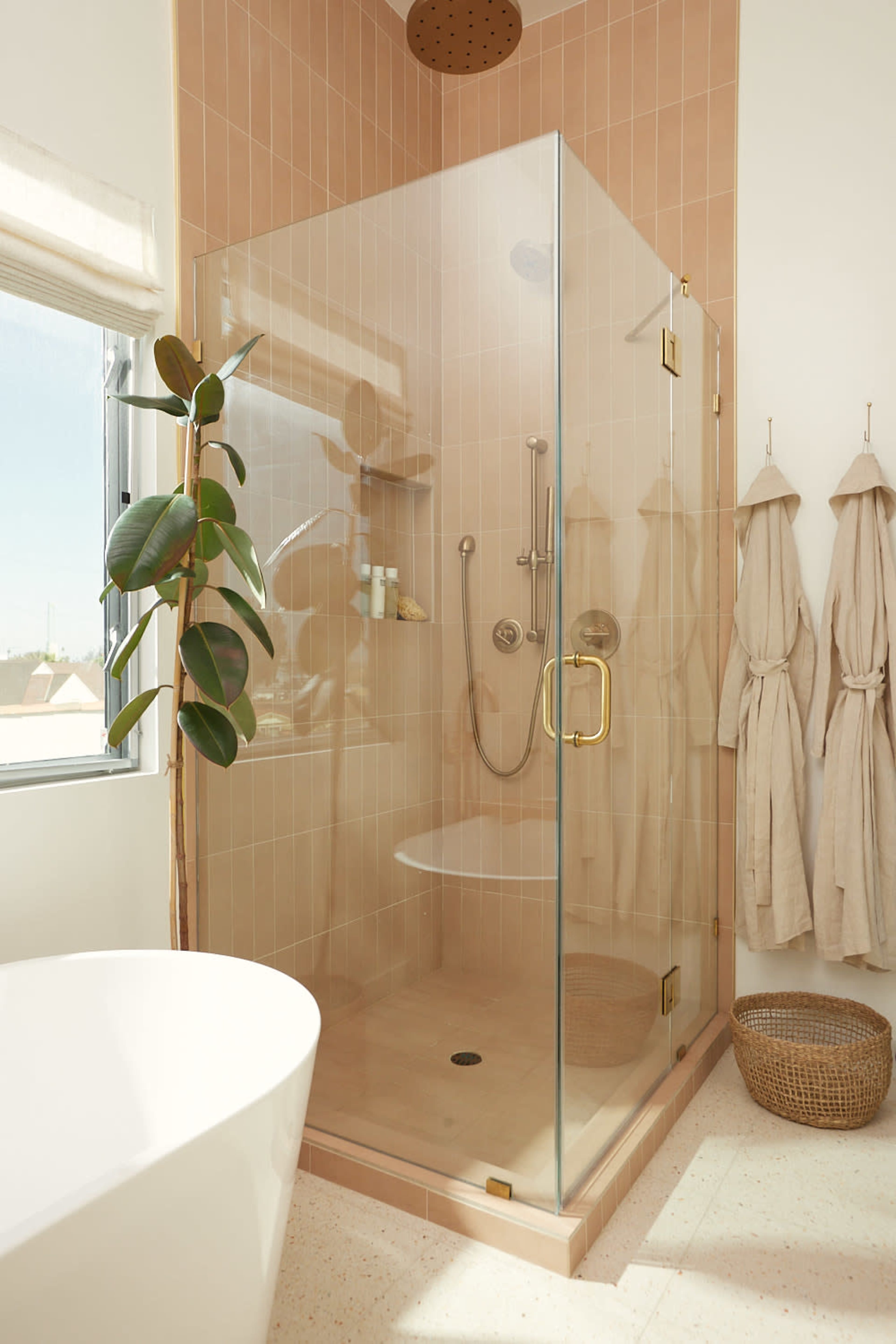 A modern bathroom featuring a glass shower enclosure, a freestanding bathtub, and beige tiled walls, with a potted plant and two hanging bathrobes nearby.