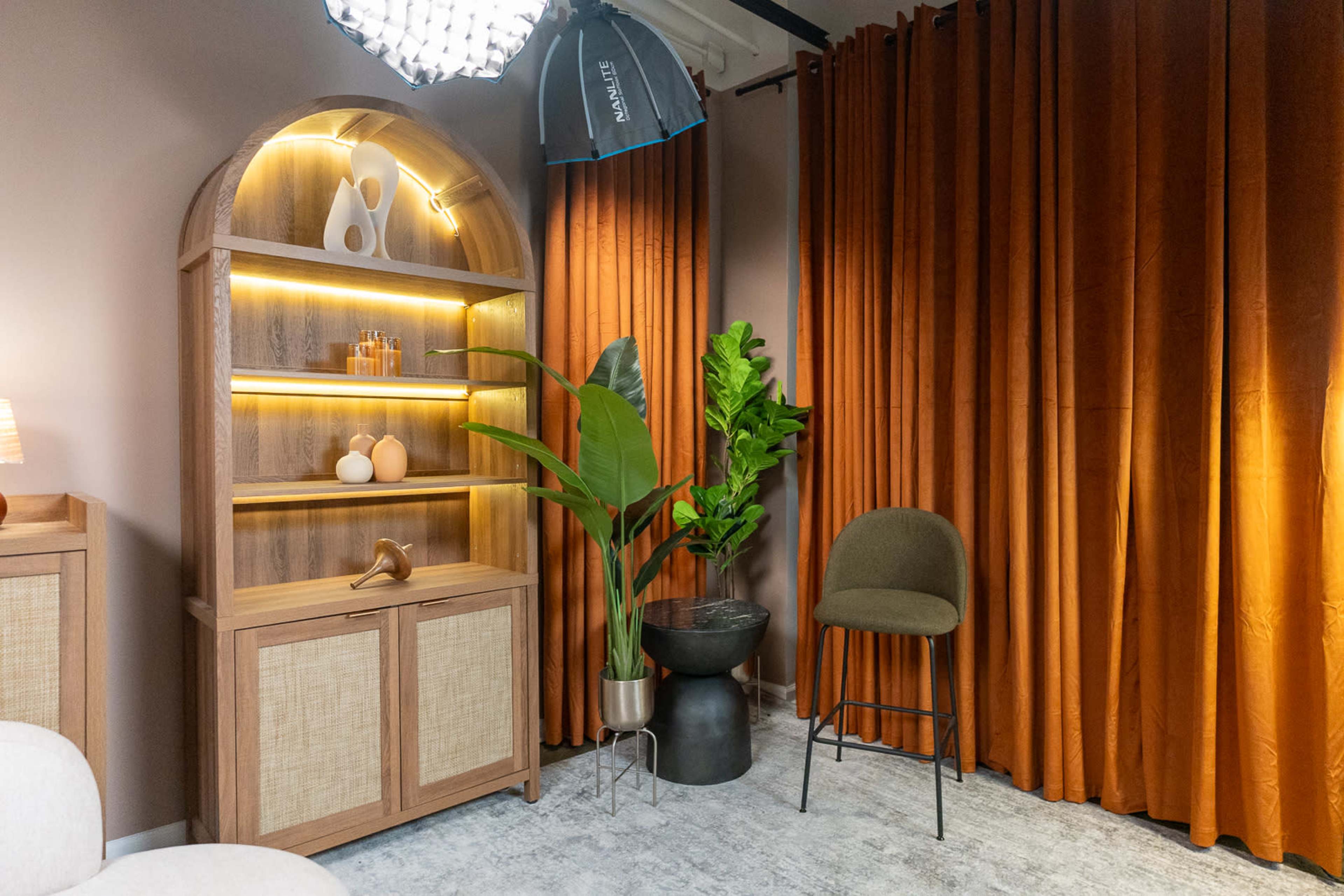 The image shows a cozy interior scene featuring a wooden shelving unit with decorative items, a large leafy plant, a stool, and elegant curtain drapery in warm tones.