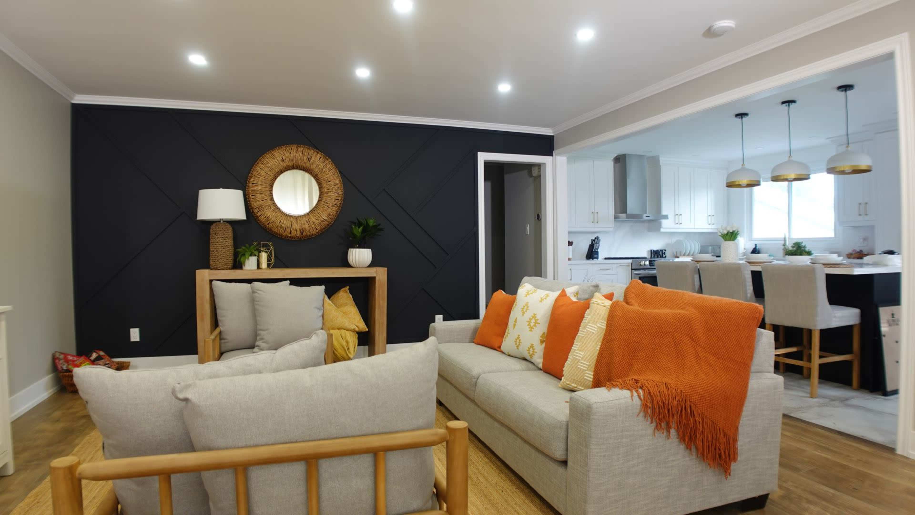 A living room features a light-colored sofa, an orange throw blanket, and a decorative wall with a round mirror, adjacent to a kitchen with white cabinetry.