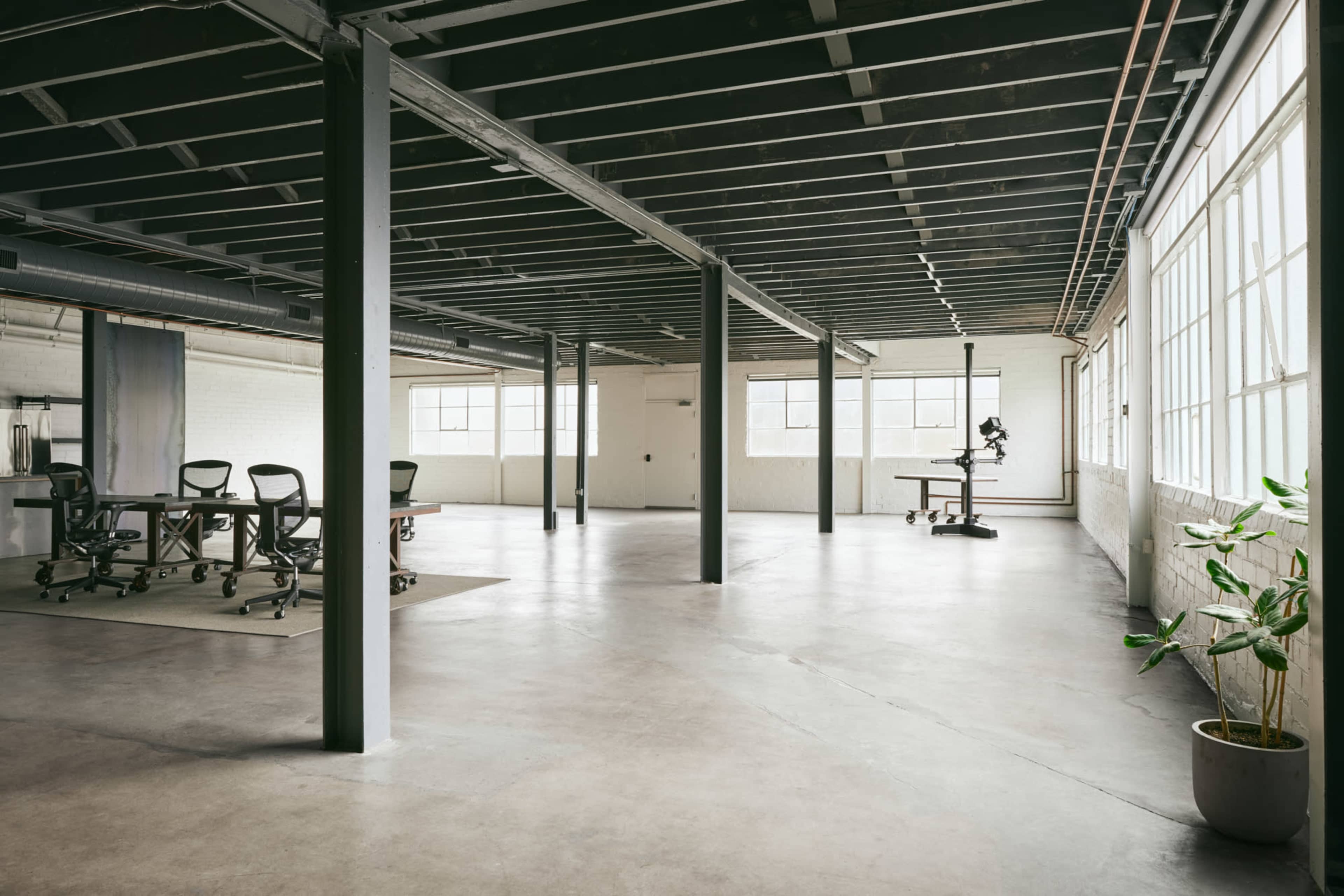 A spacious, industrial-style office with large windows, concrete floors, a meeting table with chairs, and a corner desk with an ergonomic chair.