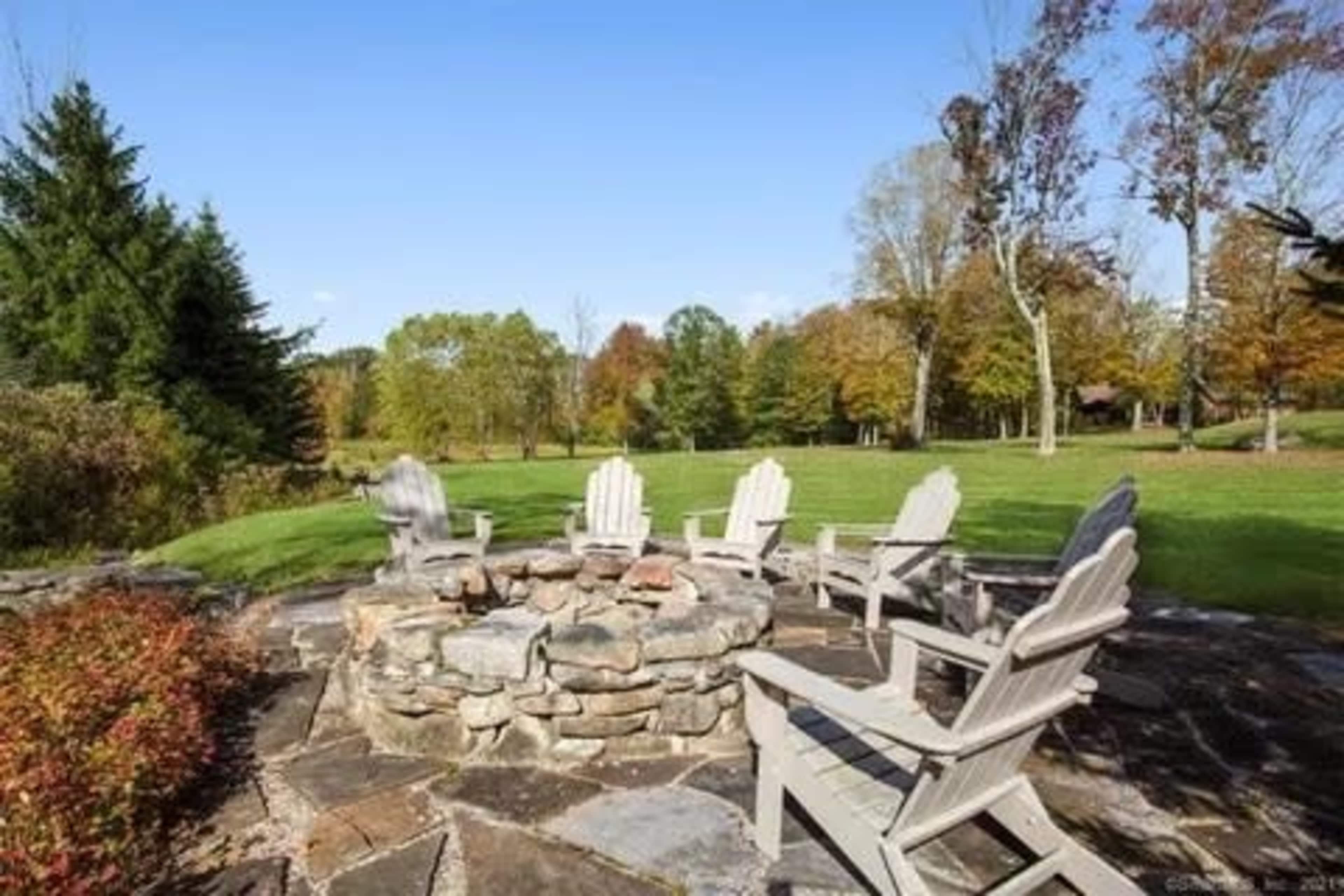 The image shows a stone fire pit surrounded by several wooden chairs in a grassy outdoor setting with trees in the background.
