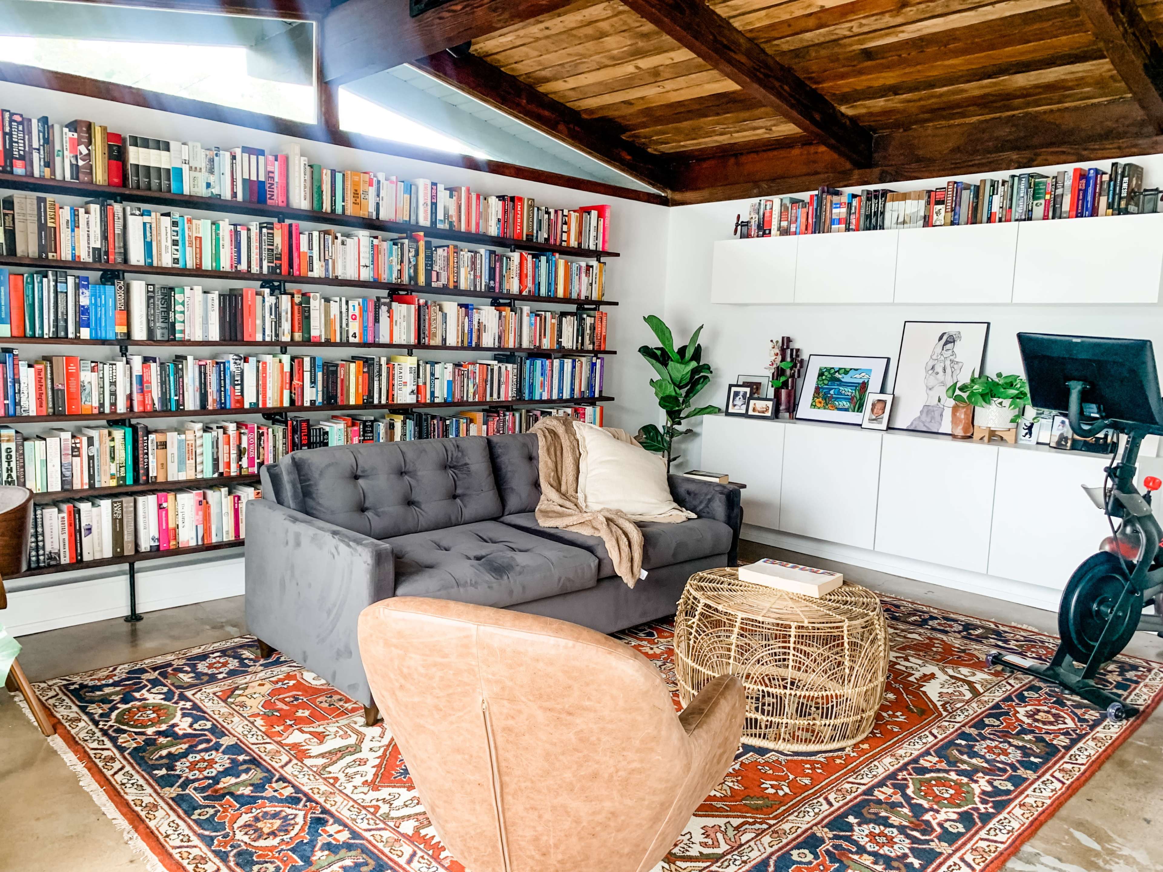 A cozy living room features a large bookshelf filled with books, a gray sofa, a round wicker coffee table, and a single leather chair on a patterned rug.