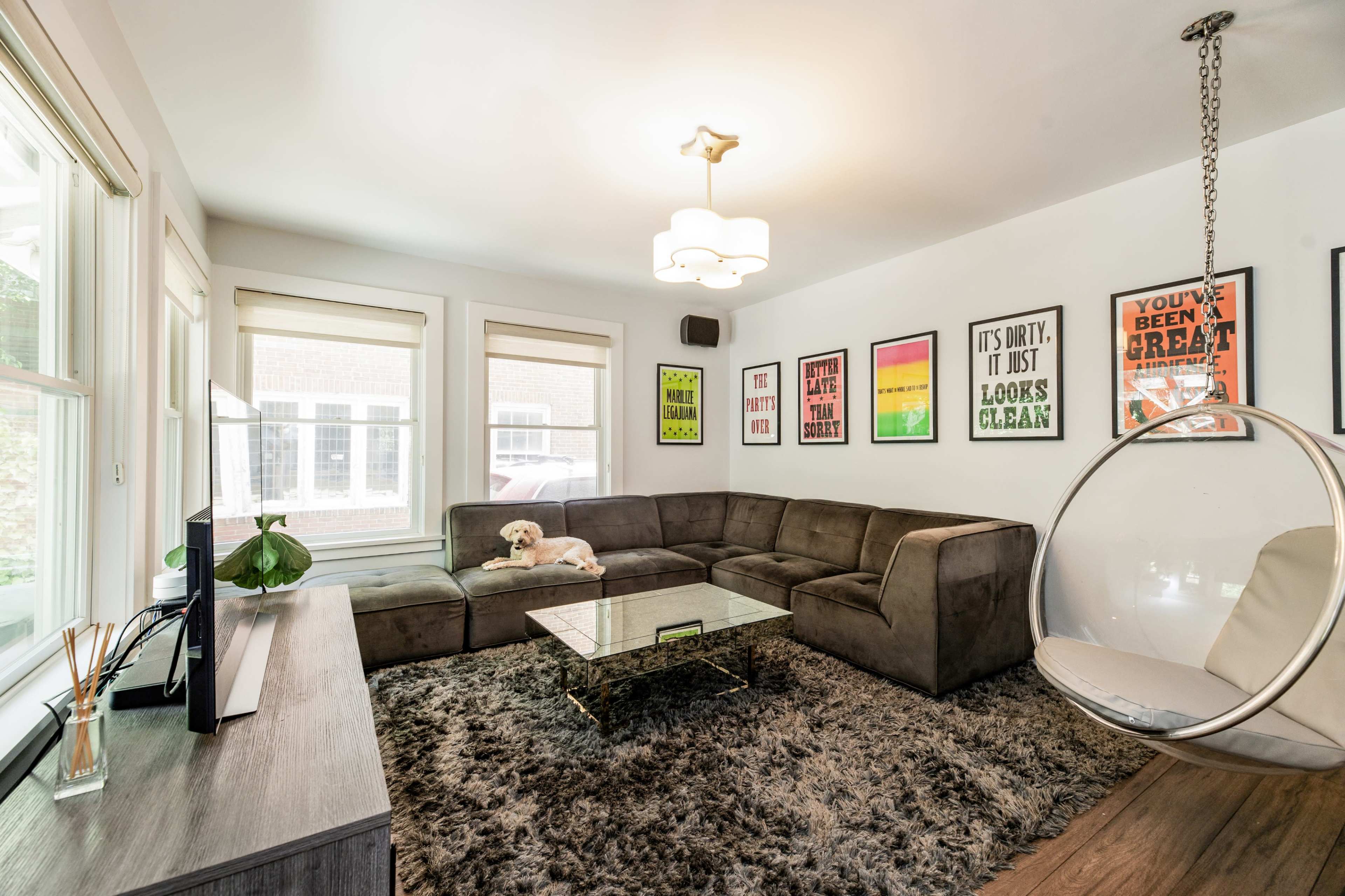 A modern living room features a sectional sofa, a glass coffee table, colorful framed posters on the walls, and a hanging chair.
