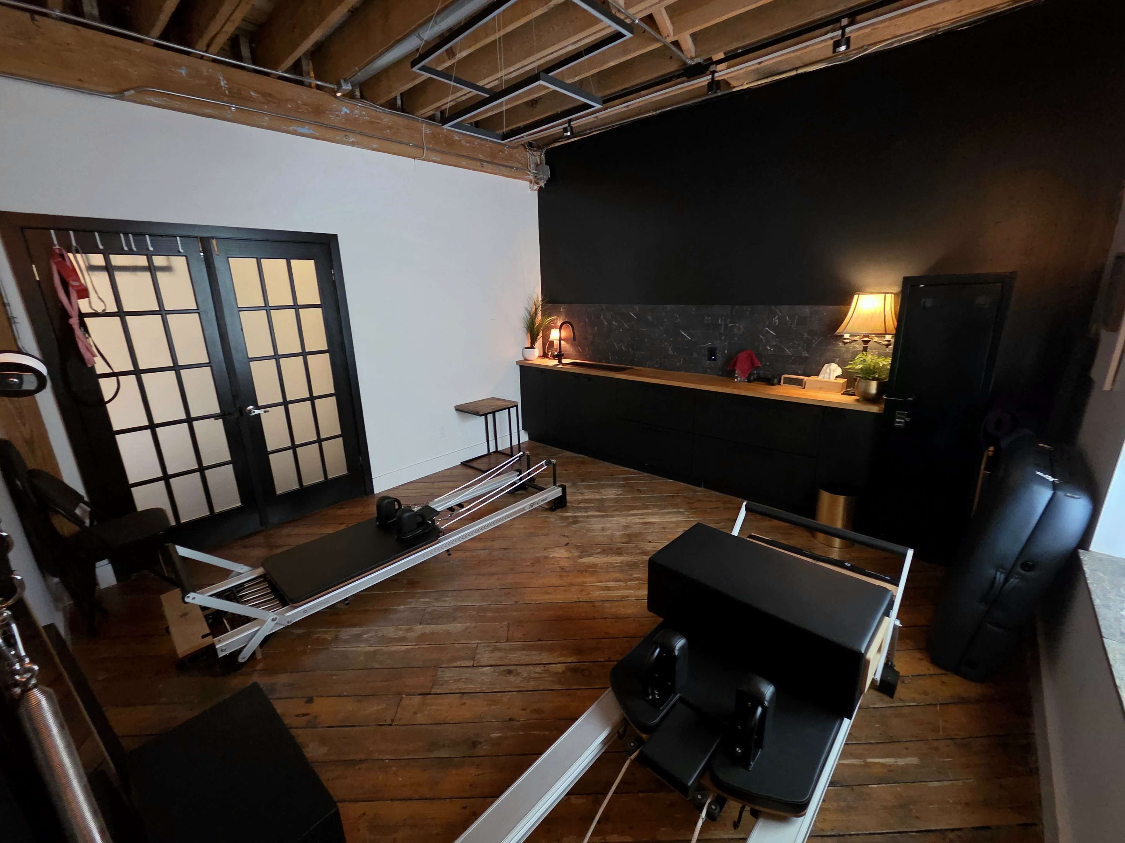 The image shows a spacious room with wooden floors, featuring a pilates reformer on one side and a reception area with a dark countertop and a lamp on the other.
