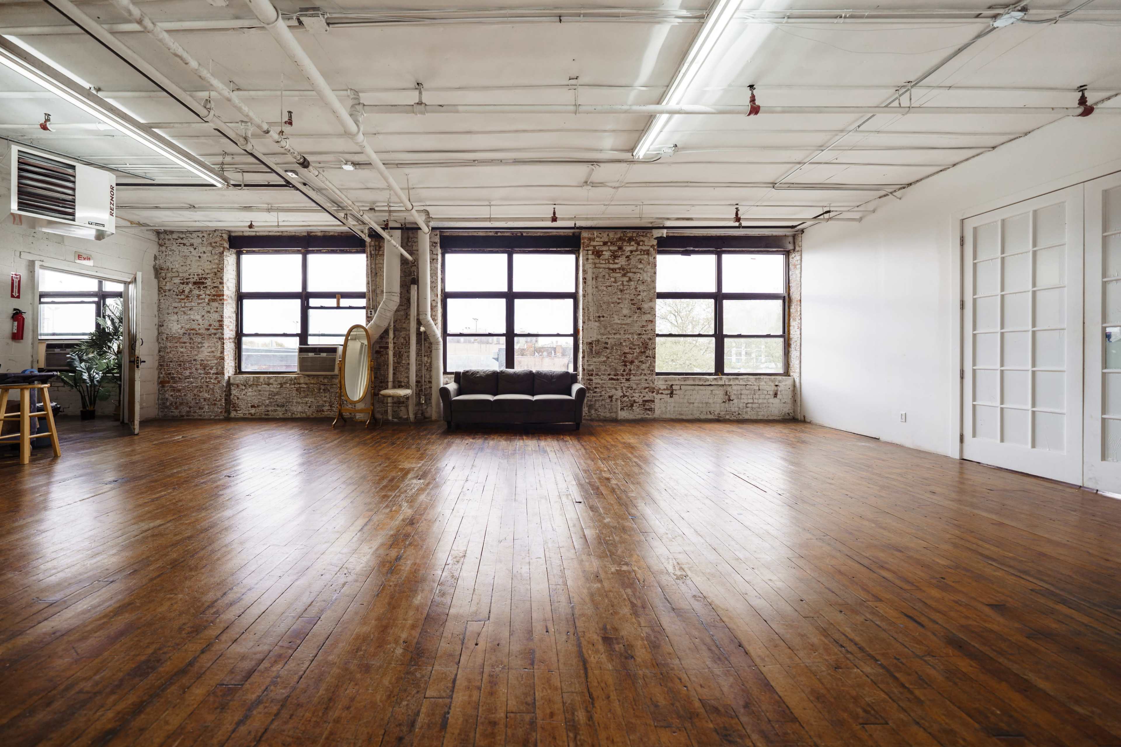 The room features a large, empty wooden floor with exposed brick walls, tall windows, and a sofa in the center.