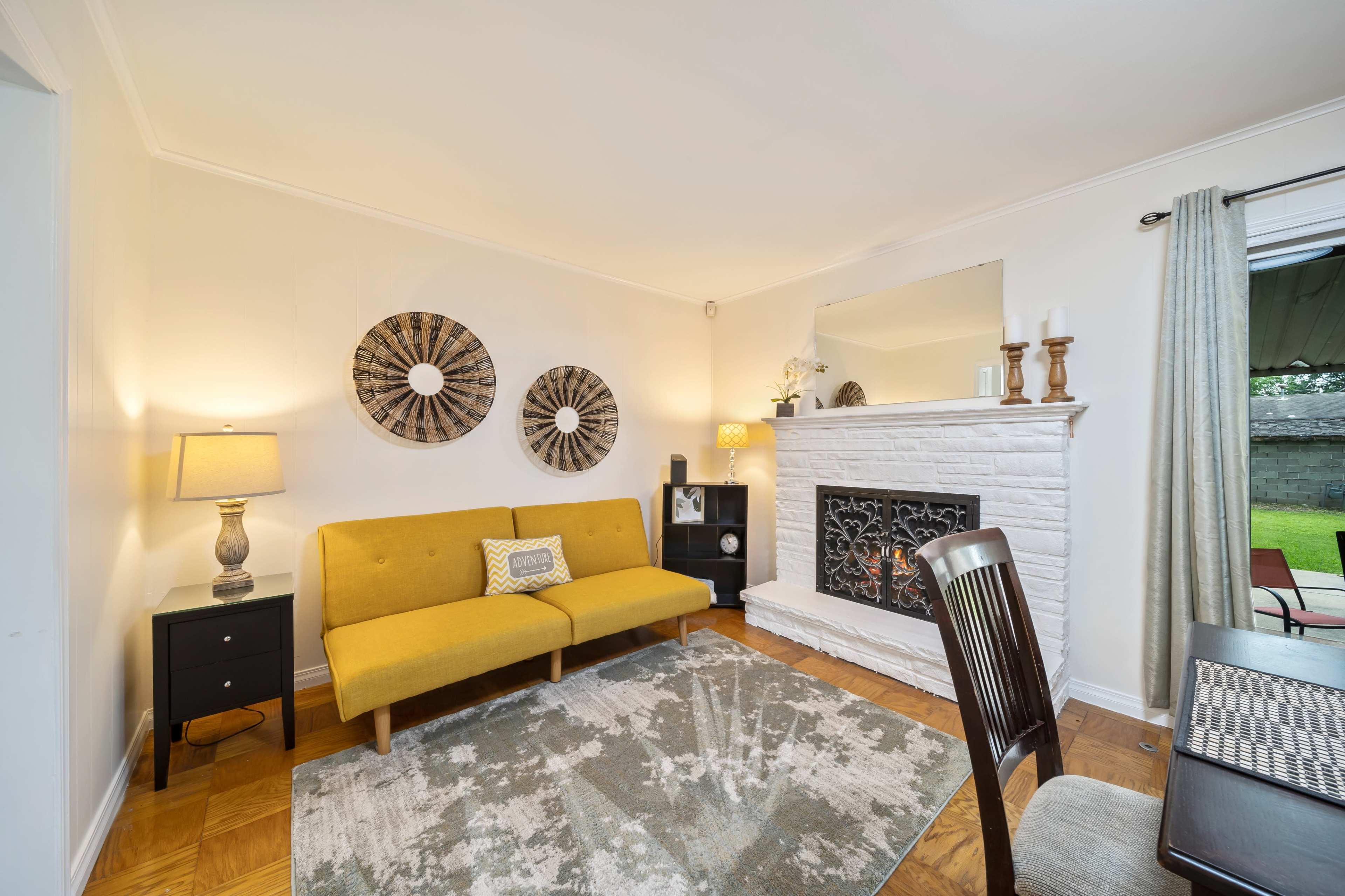 The image shows a living room with a yellow sofa, a white brick fireplace, and a small dining area featuring a wooden table and chairs.
