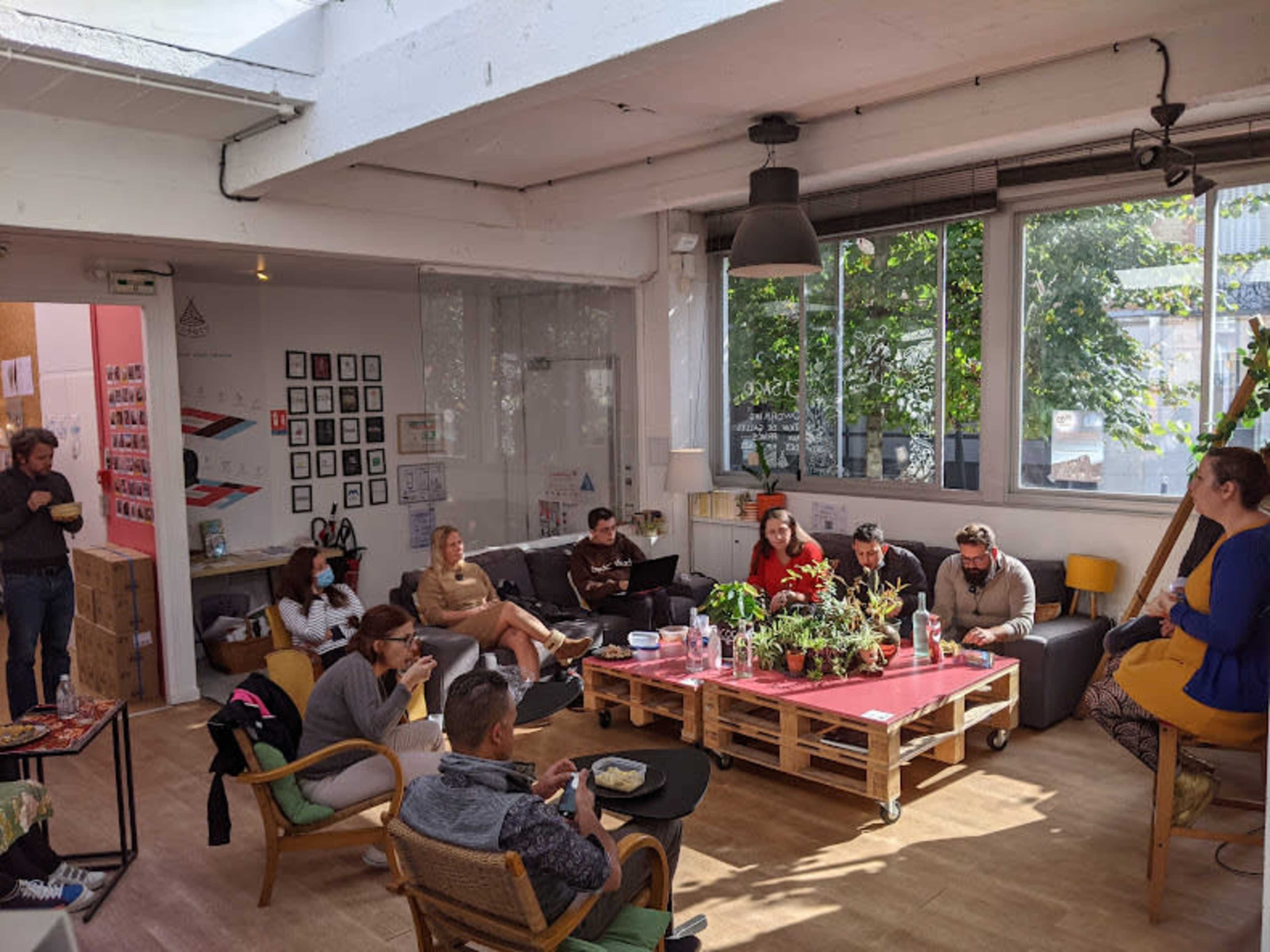 A group of people are seated in a bright, open workspace filled with plants and furniture, engaging in various activities such as conversation and work.