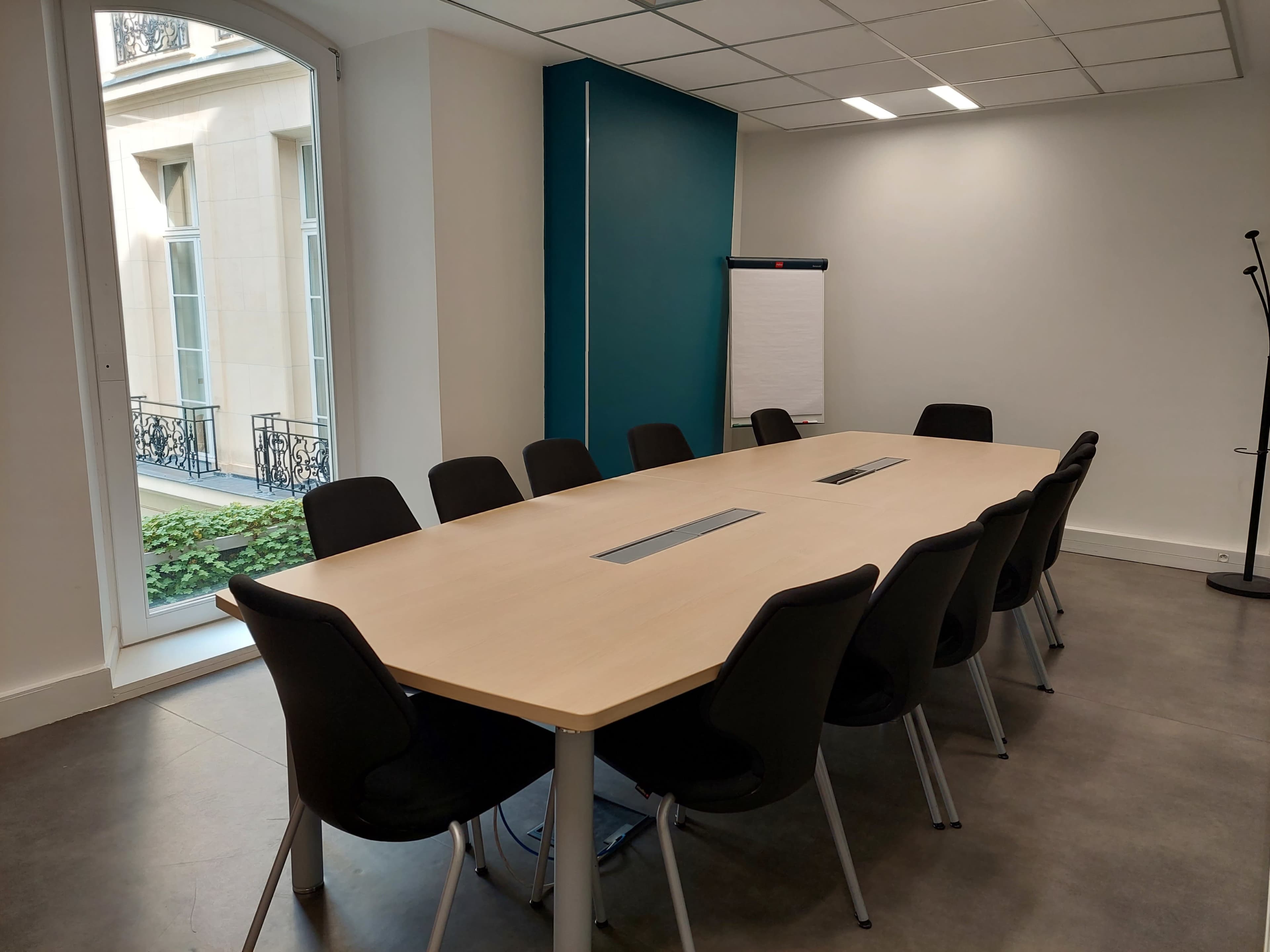 A modern conference room with a long wooden table surrounded by black chairs, featuring a flip chart and large windows letting in natural light.