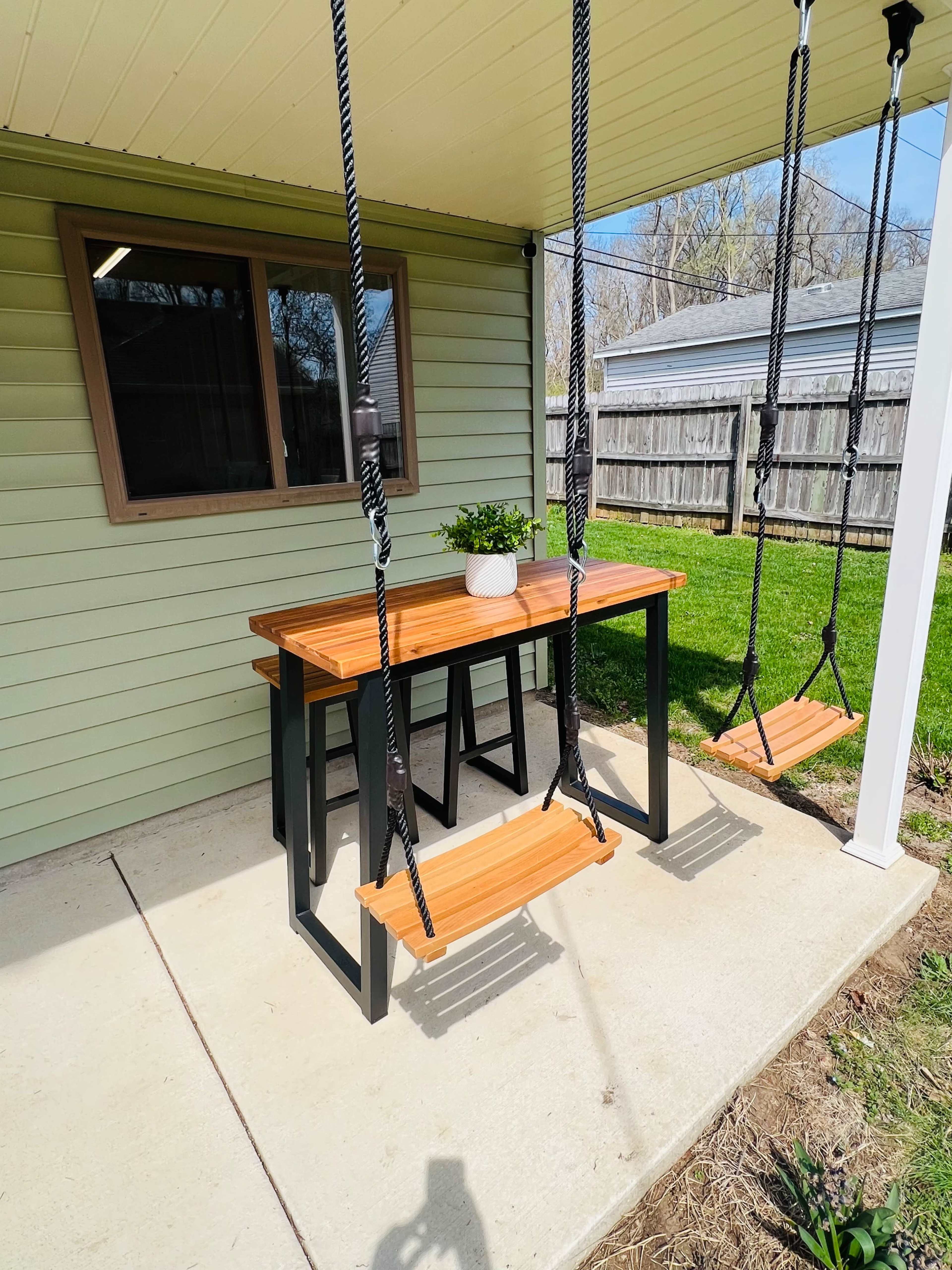 A table with two swings attached is set under a covered patio next to a grassy area.